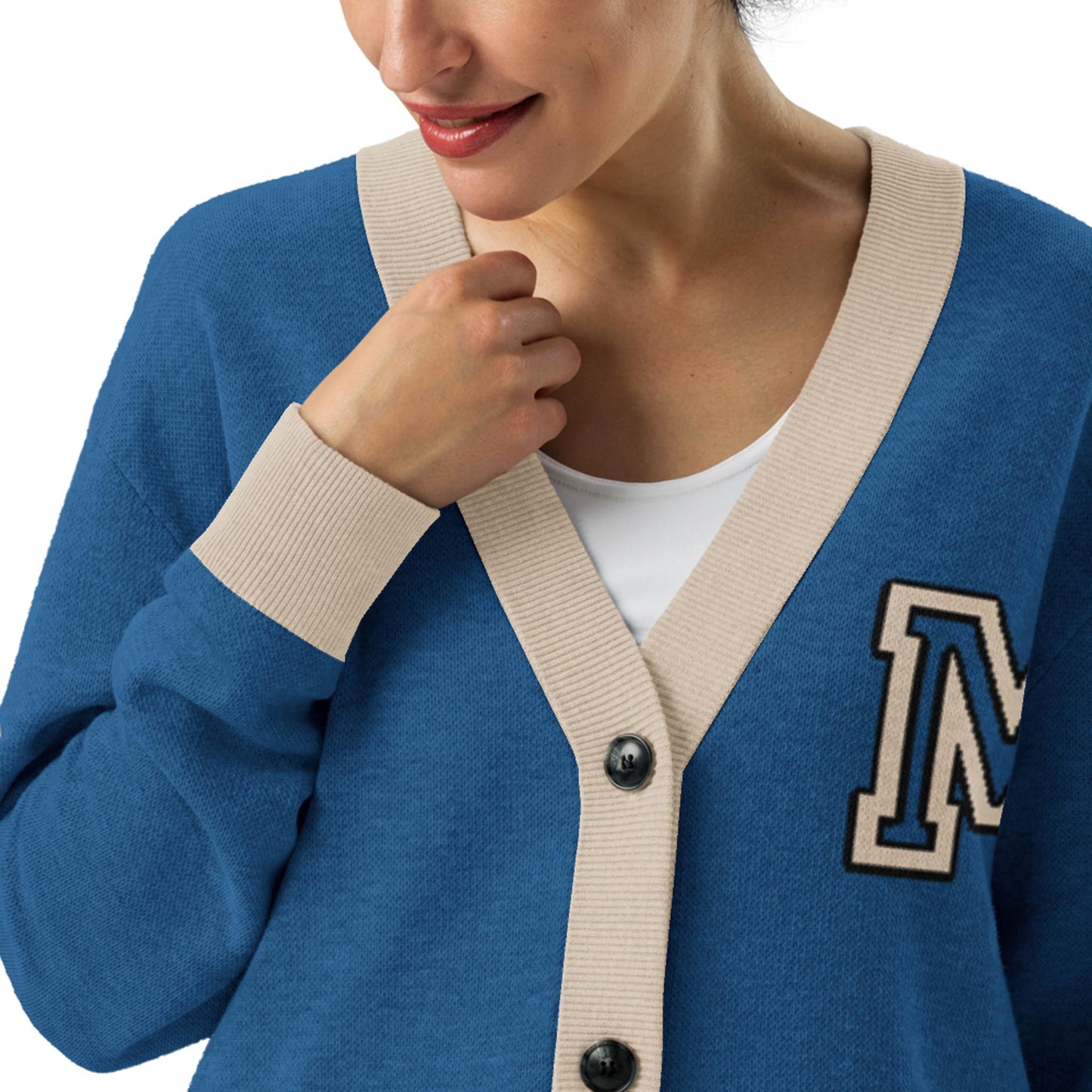 A woman wears a soft, jacquard knit blue cardigan with beige trim and an embroidered letter M on the chest, over a white top. She is smiling slightly and has one hand near her collar.