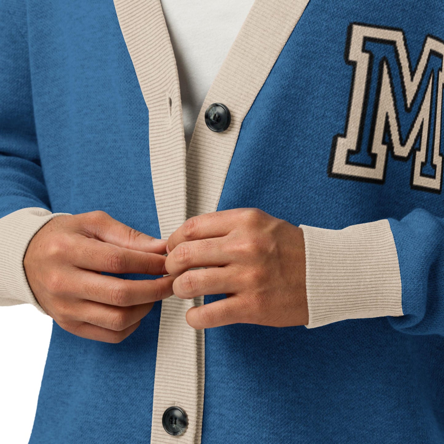 A person buttoning a soft, blue unisex cardigan with beige trim and large buttons. The jacquard knit cardigan features a white letter M patch on the left side. Only the persons hands and torso are visible.
