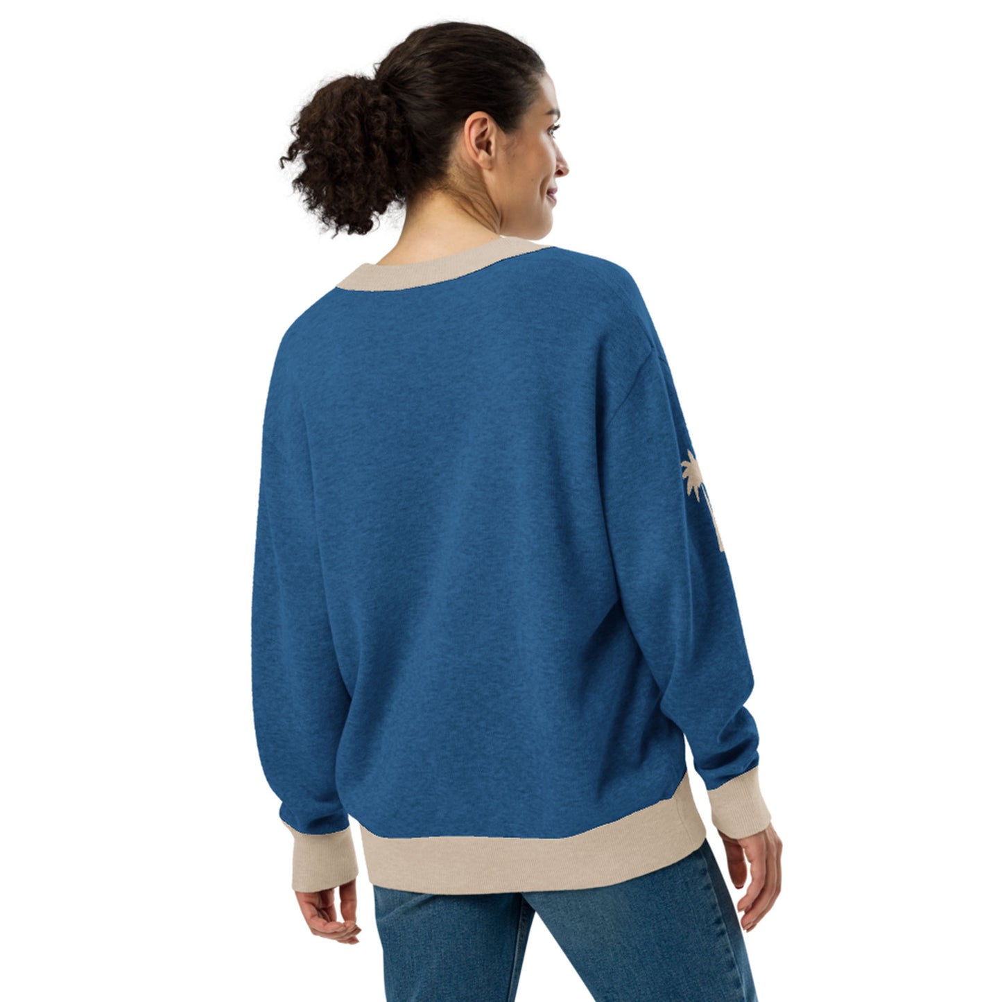 A woman with curly hair in a ponytail is standing and smiling, wearing a loose two-tone cardigan with beige cuffs and hem, and blue jeans. She is facing away from the camera.