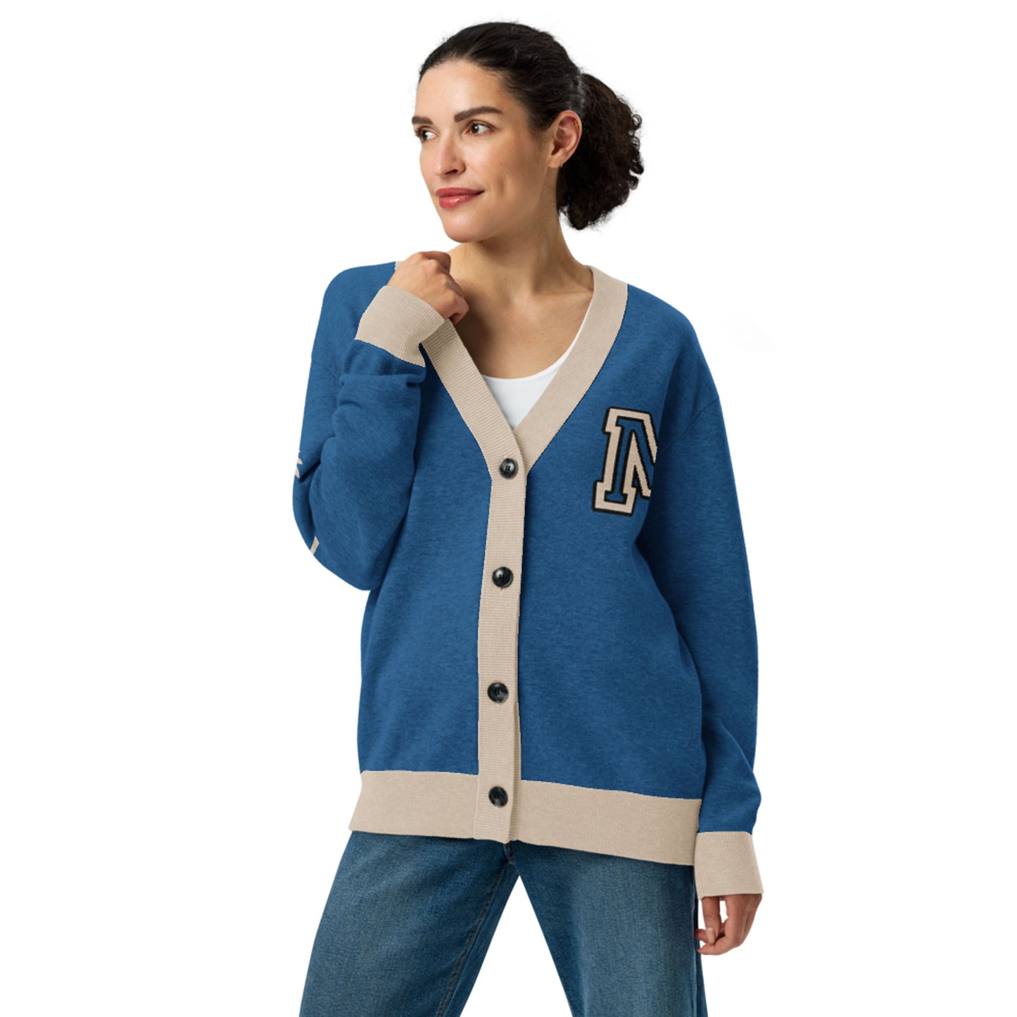 A woman wearing a blue two-tone cardigan with beige trim and an A patch over a white top and blue jeans stands facing slightly left, with one hand touching the collar and a slight smile on her face.