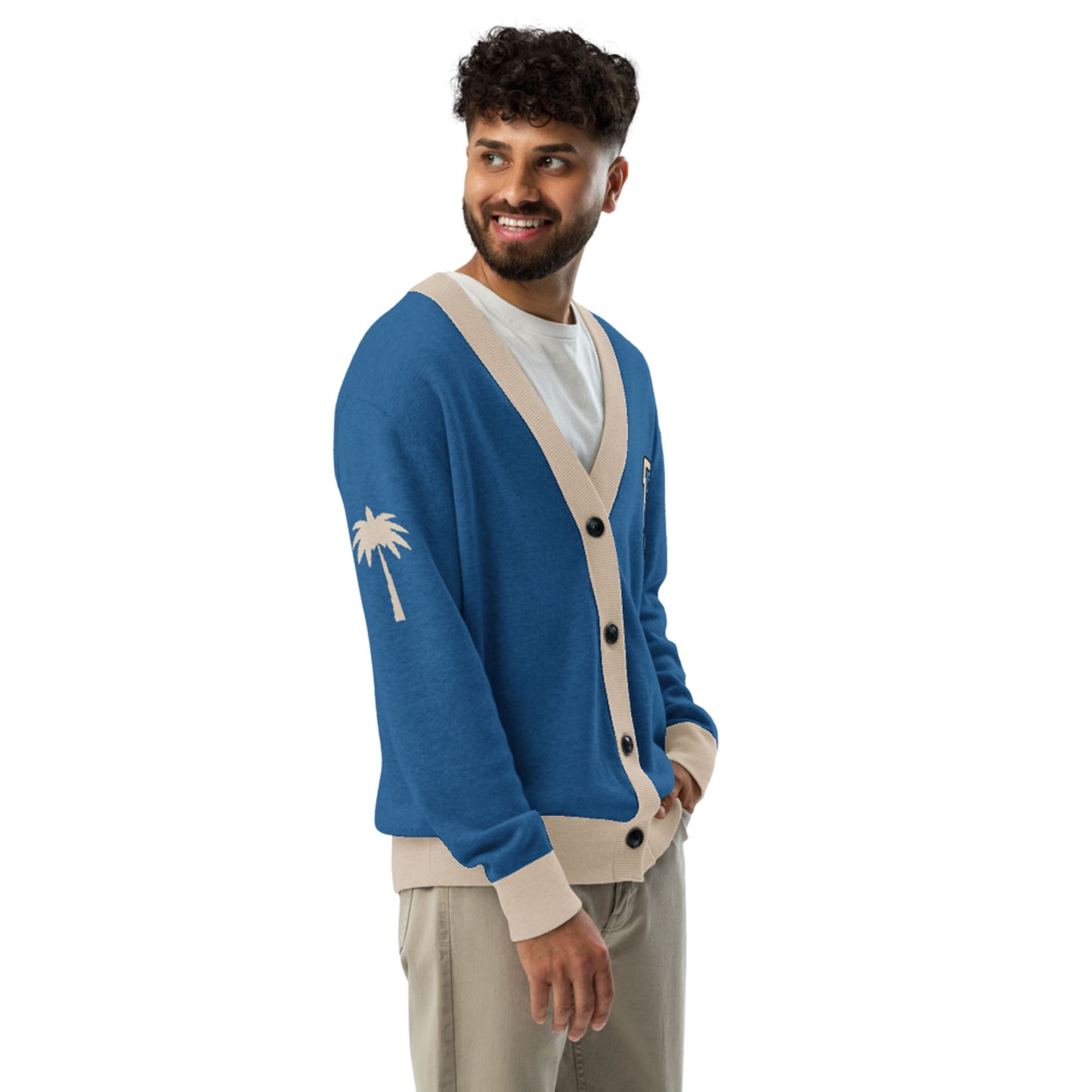 A smiling man with curly hair and a beard wears a blue and beige two-tone cardigan featuring a palm tree jacquard knit design on the sleeve, paired with beige pants, standing against a plain white background.