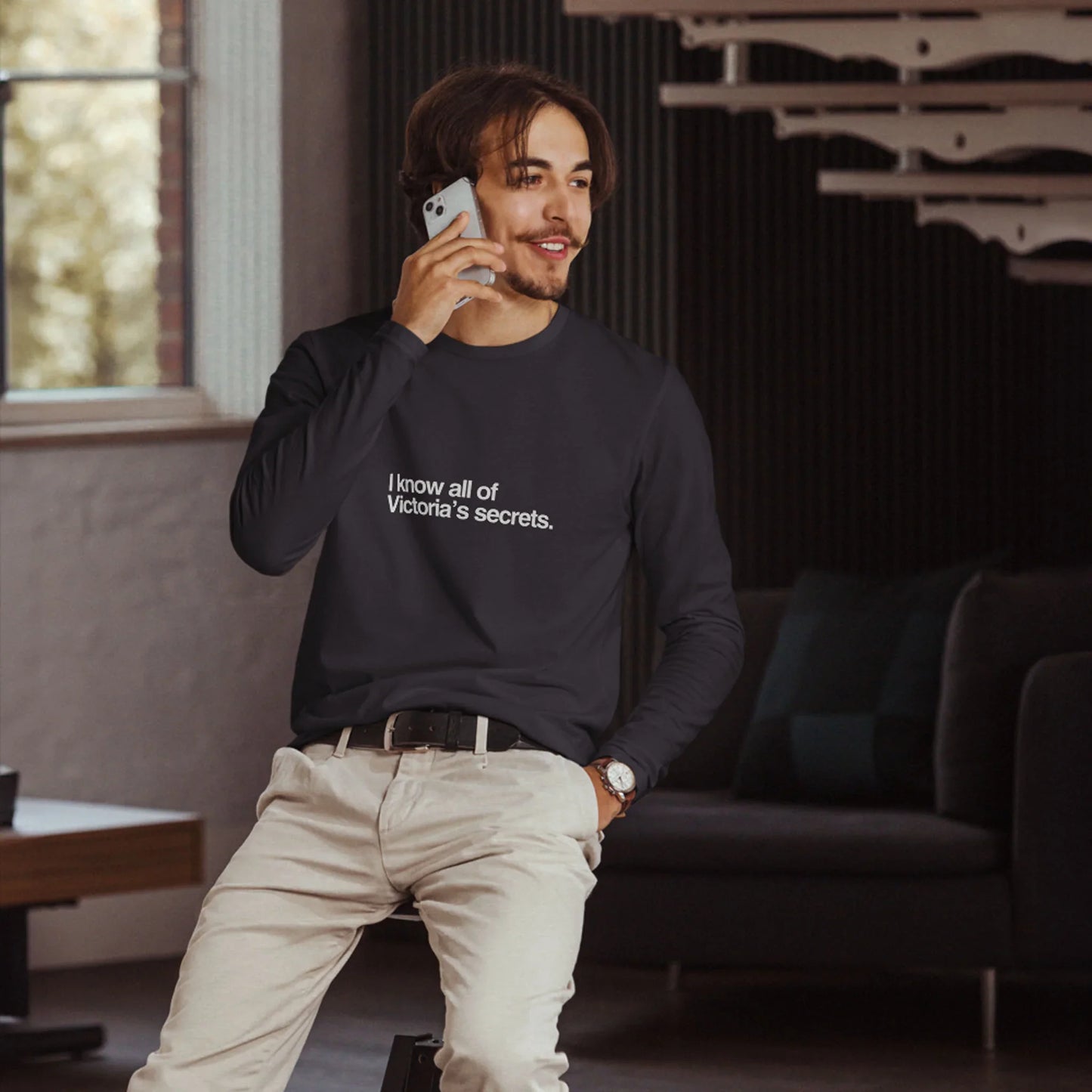 Man wearing a black long-sleeve graphic t-shirt with text, standing in a room with dark walls and a window.