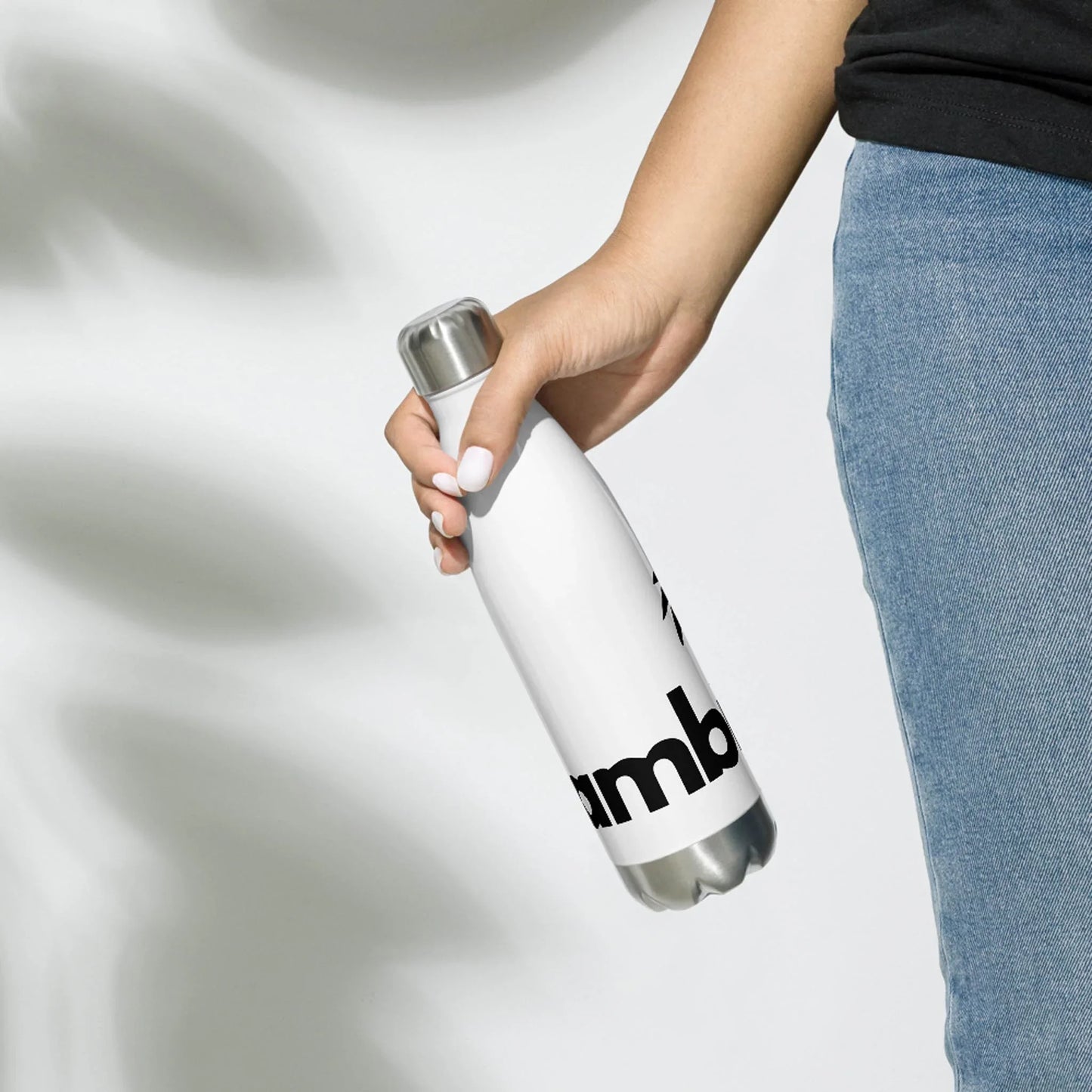 A person in blue jeans and a black shirt holds the Printful Pura Stainless Water Bottle, 17oz, in white with a silver cap and black text, against a plain light background.