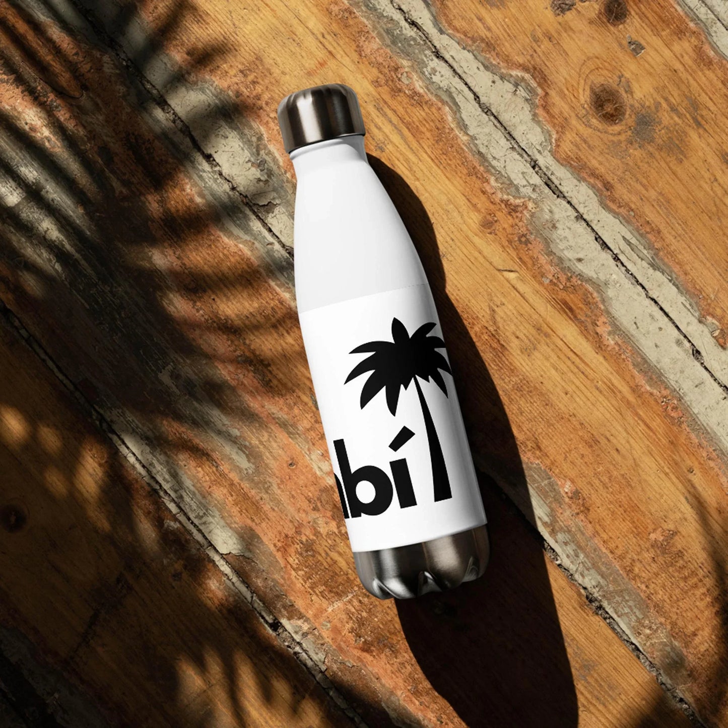The Pura Stainless Water Bottle by Printful, 17oz in white with a black palm tree design and partial text, rests on a wooden surface with palm leaf shadows cast across the scene.