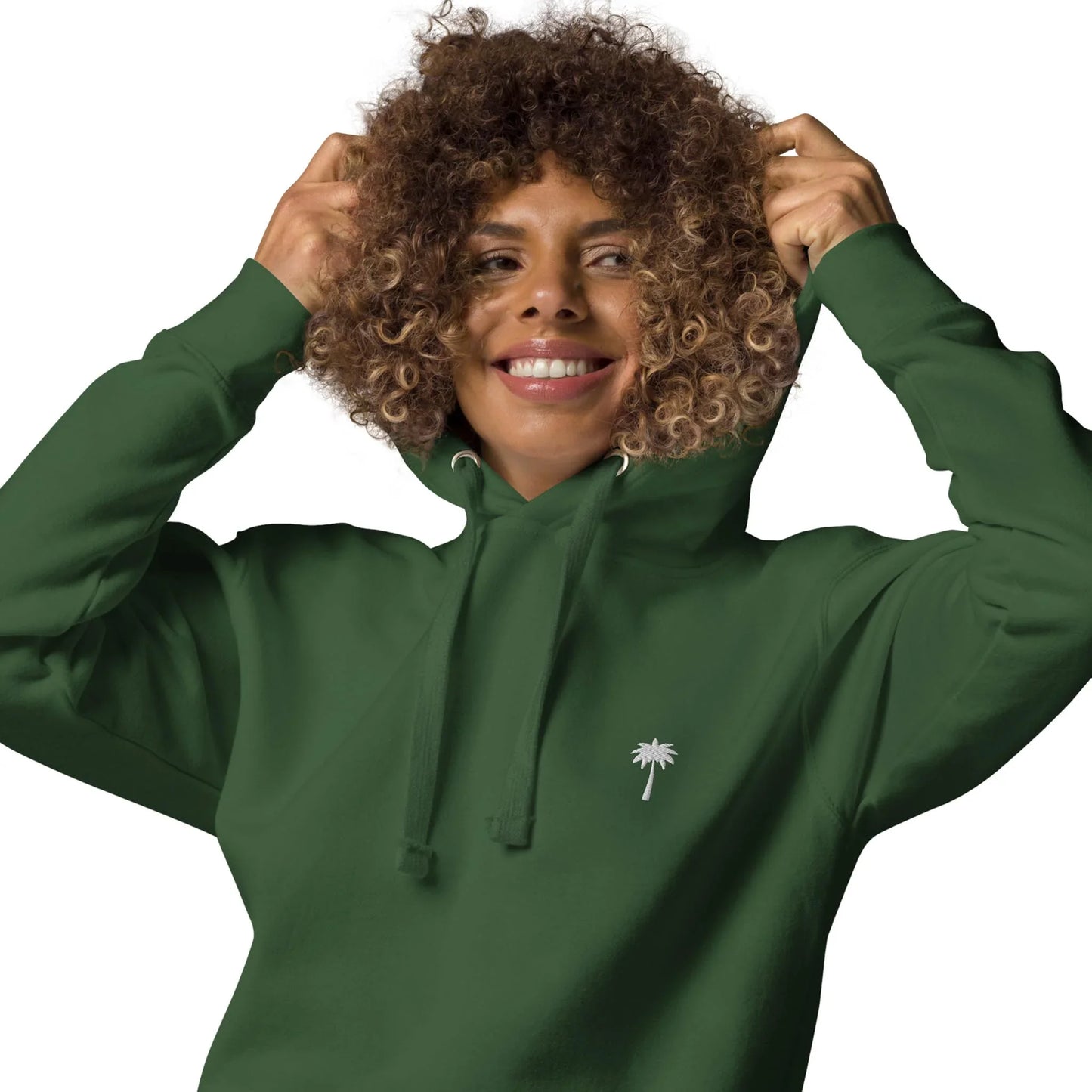 A smiling person with curly hair wears a dark green Mambí hoodie with a small white palm tree logo on the chest and holds the hood with both hands against a white background.