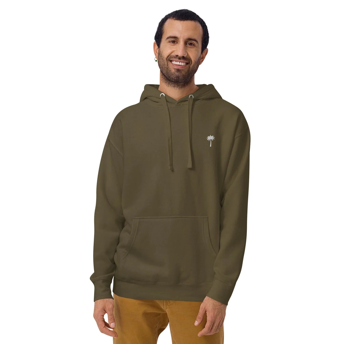 A man with dark hair and a beard is smiling, wearing an olive green Mambí hoodie with a small palm tree graphic on the chest and tan pants, standing against a plain white background.