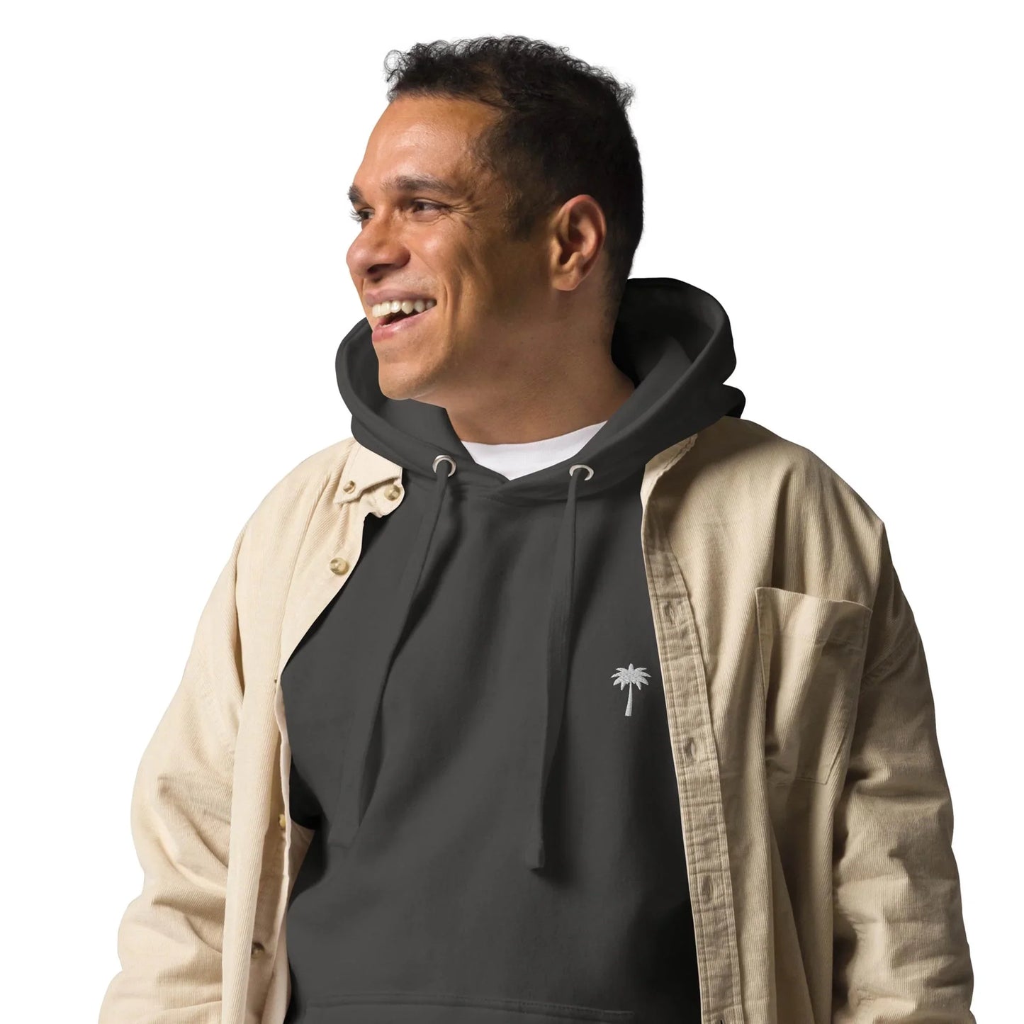 A smiling man wears a Mambí black hoodie with a small white palm tree design and a beige button-up shirt over it, standing against a plain white background.