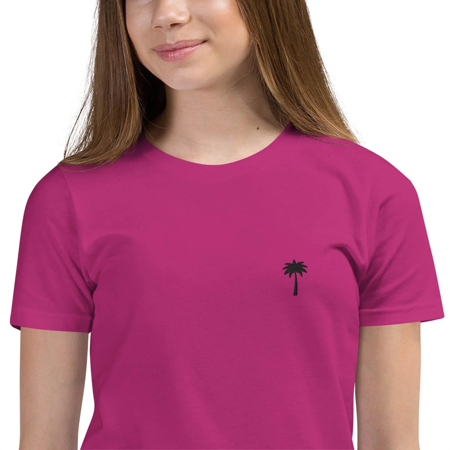 A young person wearing a bright magenta Mambí t-shirt with a small black palm tree graphic on the left chest, smiling softly. Only the upper half of the body is visible.