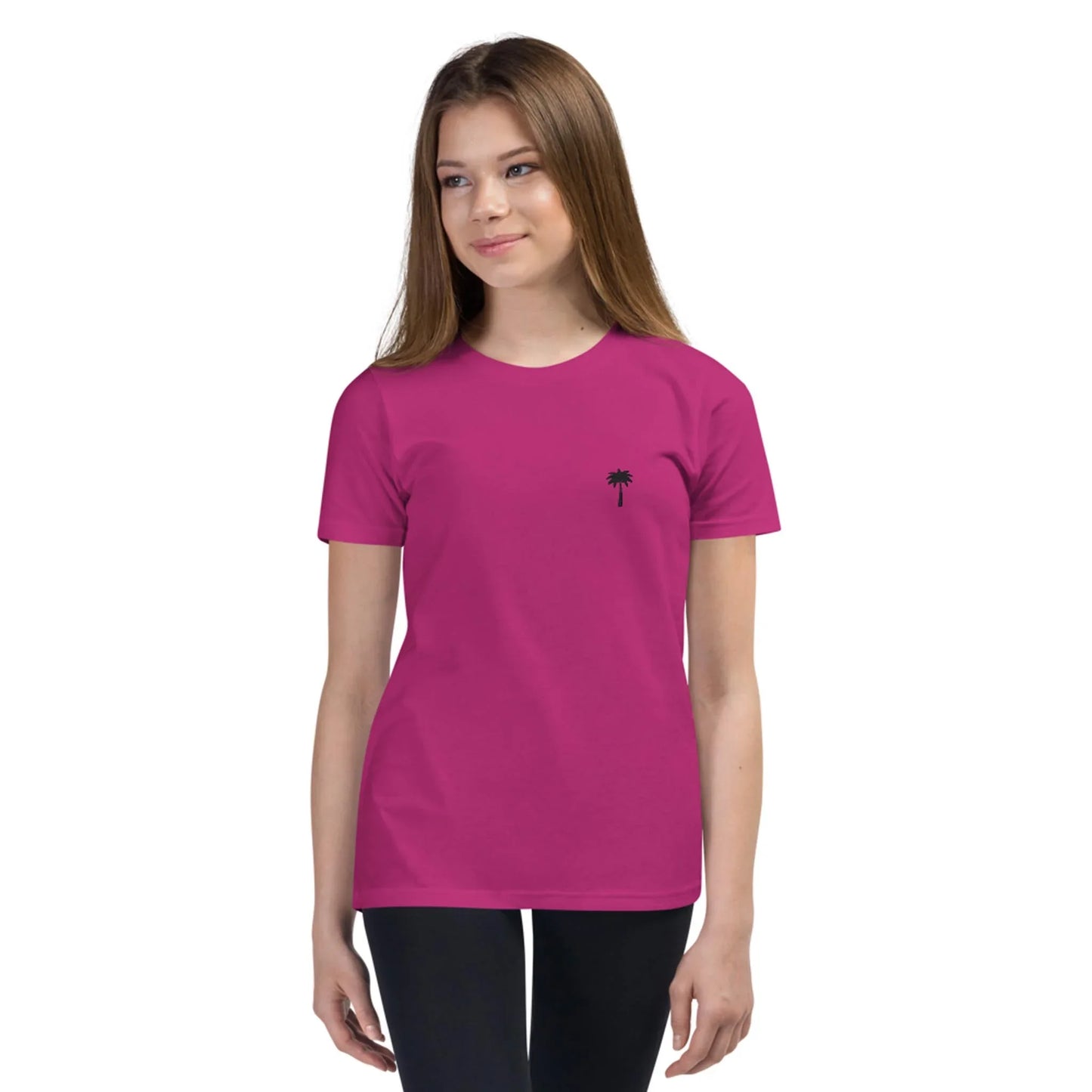 A young woman with long brown hair wears a bright pink Mambí t-shirt featuring a small black palm tree graphic on the left chest. She is also wearing black pants and stands facing slightly to the side, smiling gently.