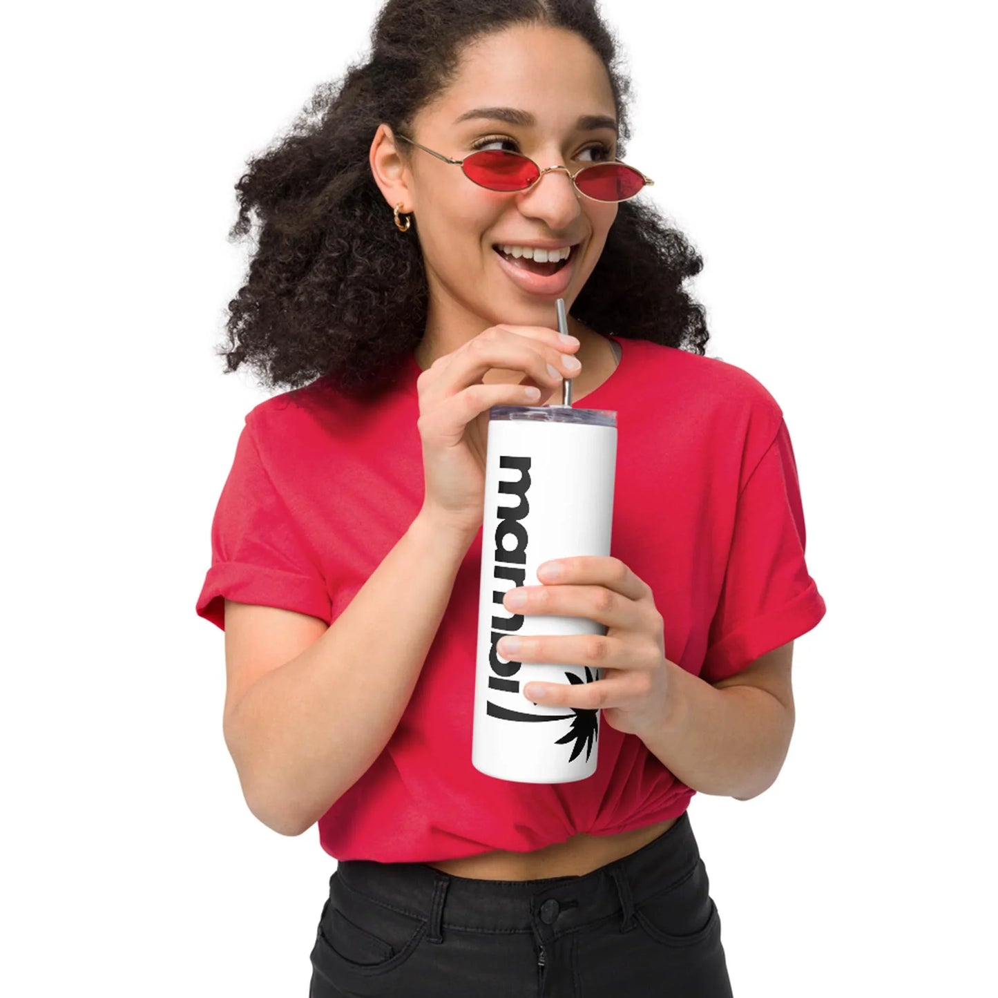 A woman with curly hair wearing red sunglasses and a red shirt drinks from a white Mambí tumbler with black text and a palm tree graphic. She is smiling and standing against a white background.