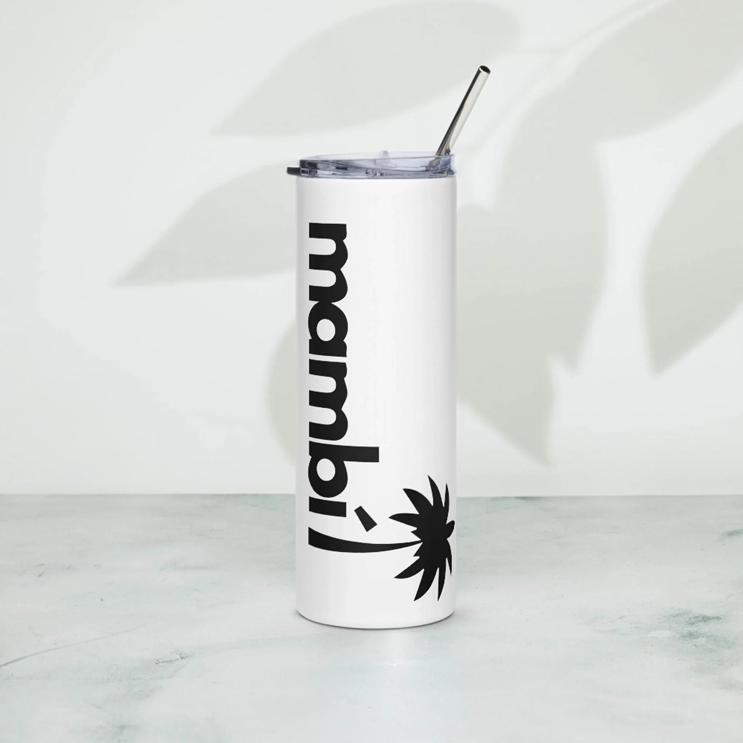 A tall white tumbler with a metal straw and clear lid, featuring the word Mambí in bold black letters and a palm tree graphic, stands on a light surface with a leaf shadow in the background.