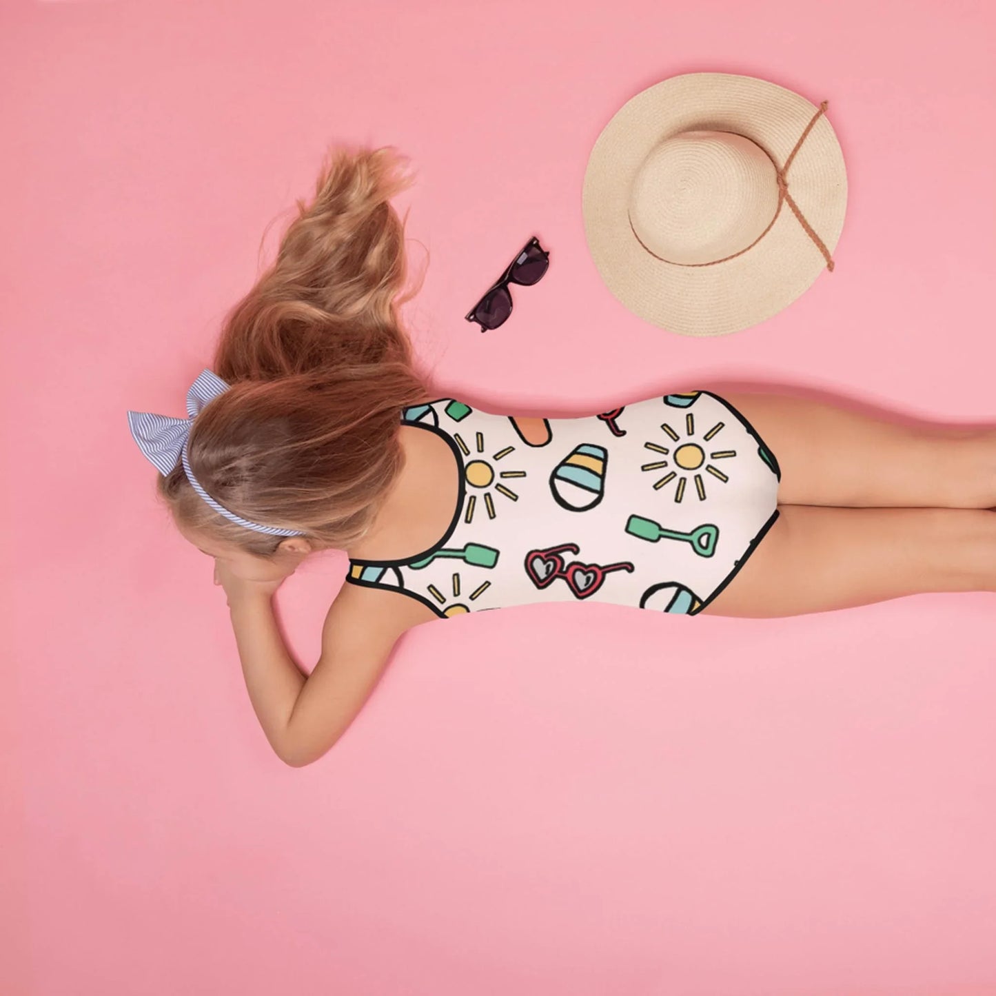 A young girl with long blonde hair, wearing a patterned swimsuit and a blue bow, lies on a pink background near a straw hat, sunglasses, and a Mambí accessory.