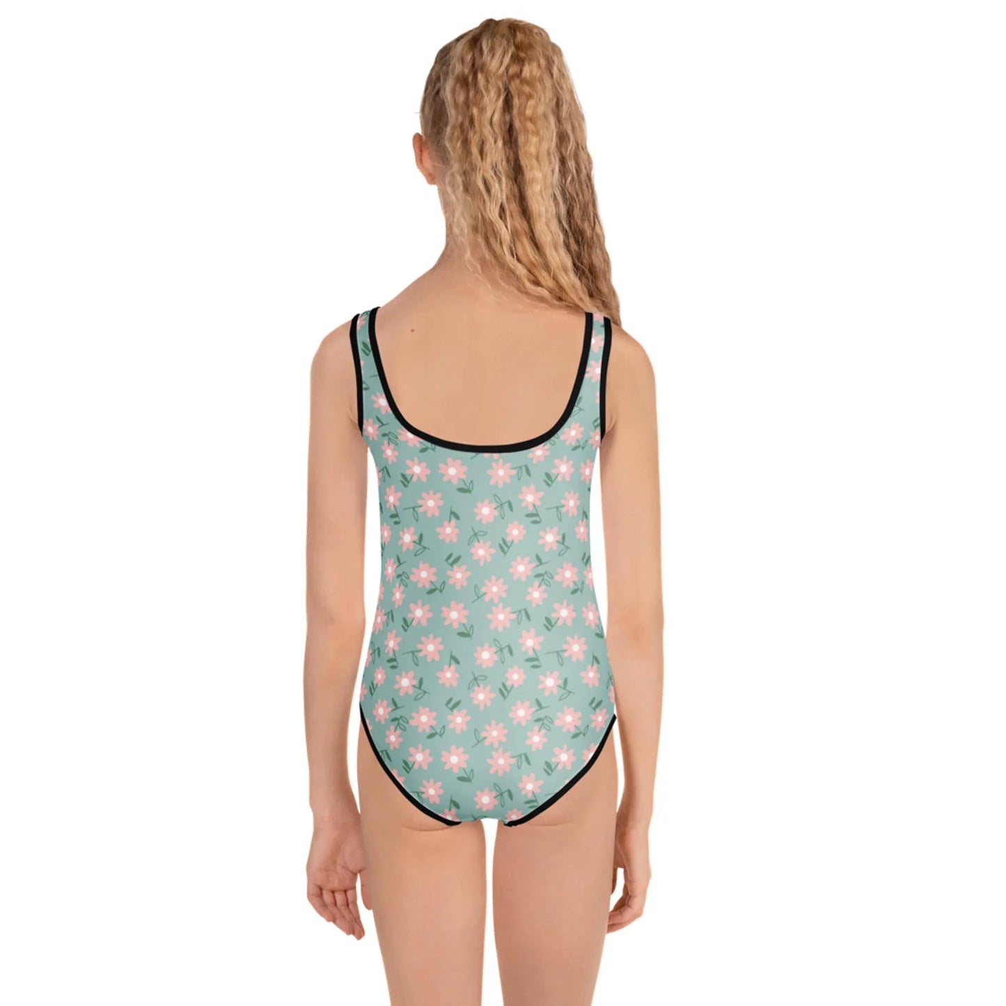 A girl with long, wavy blonde hair faces away from the camera, modeling the Printful Sara One-Piece Swimsuit for toddlers and little kids in light green with bright pink and white daisy prints and black trim against a plain white background.