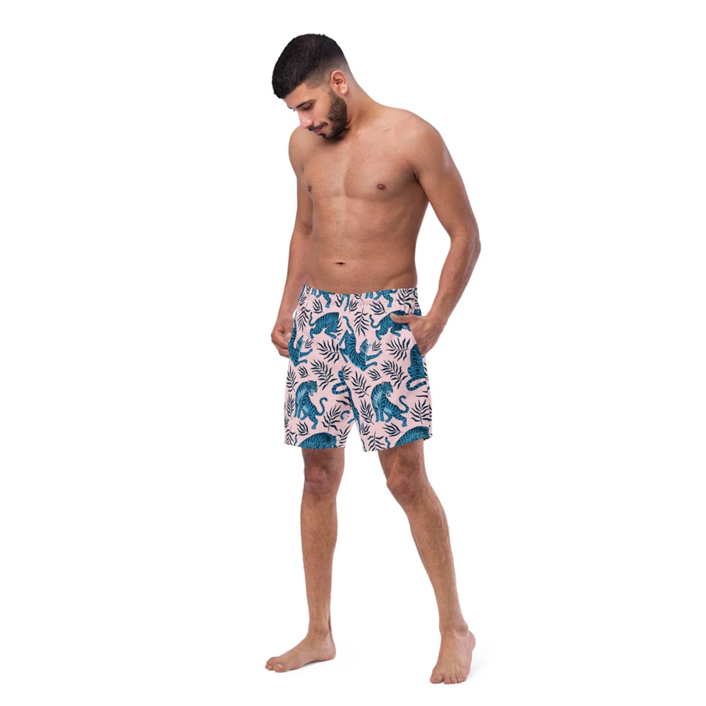 A shirtless man with a beard stands barefoot, wearing Mambí pink swim shorts with a blue tropical leaf pattern, looking down with his hands in his pockets against a white background.