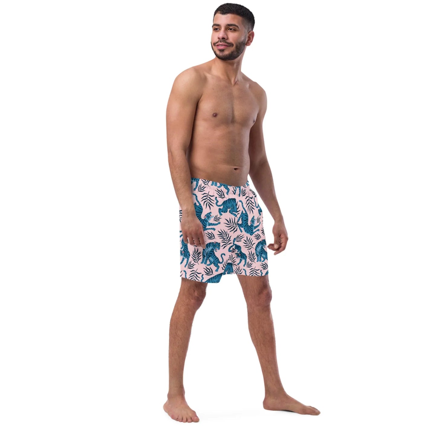 A smiling man stands barefoot against a white background, wearing Mambí swim shorts with a dark blue jungle animal and foliage print. He is shirtless and looking slightly to his right.