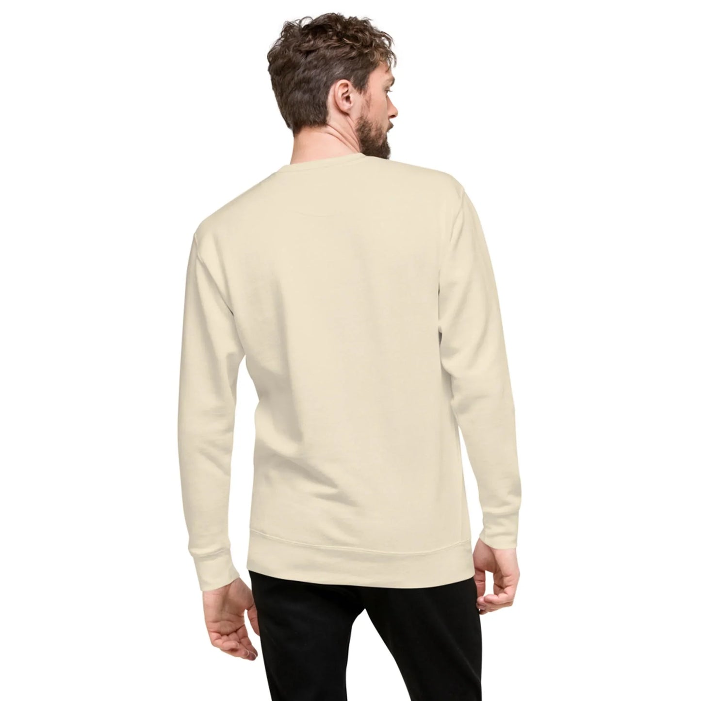 A man with short, curly hair is standing and facing away, wearing a plain, light beige Mambí sweatshirt and black pants against a white background.
