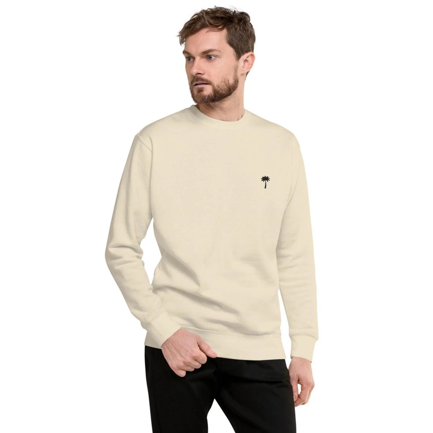 A man wearing a light beige Mambí sweatshirt with a small black palm tree graphic on the left chest stands with one hand in his pocket, looking off to the side. He has short hair, a beard, and is dressed in black pants.
