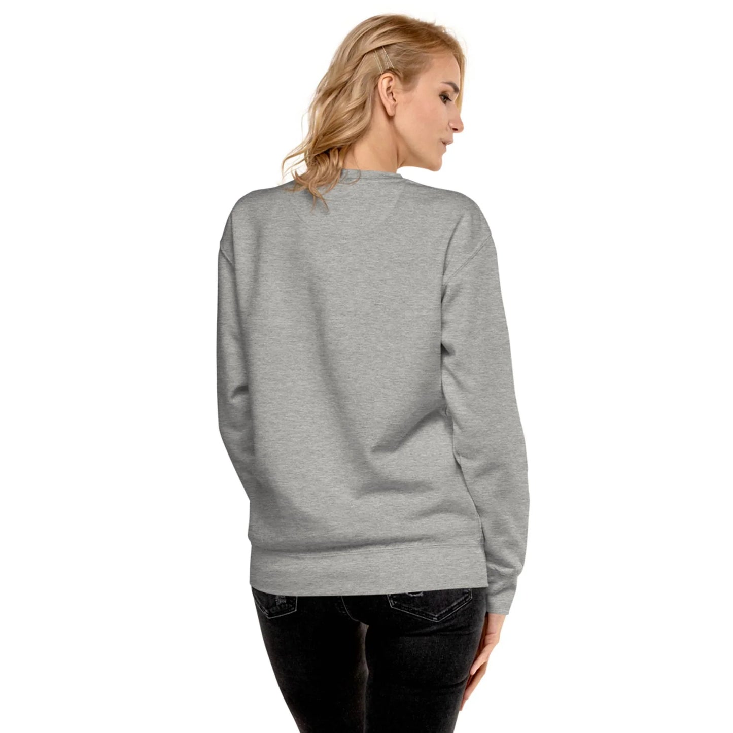 A woman with blonde hair wearing a light gray Mambí sweatshirt and black jeans stands facing away, looking over her left shoulder against a white background.