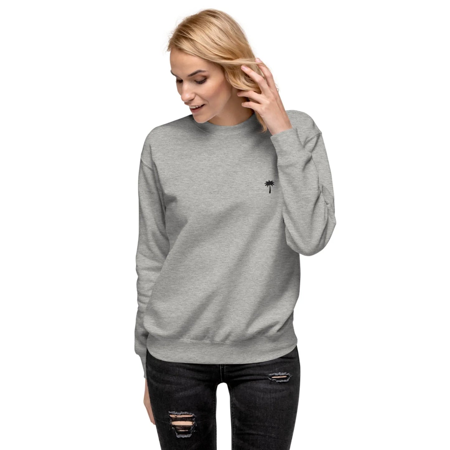 A woman wearing a light gray Mambí sweatshirt with a small black palm tree logo on the chest stands against a white background, touching her hair and smiling. She pairs it with black ripped jeans.
