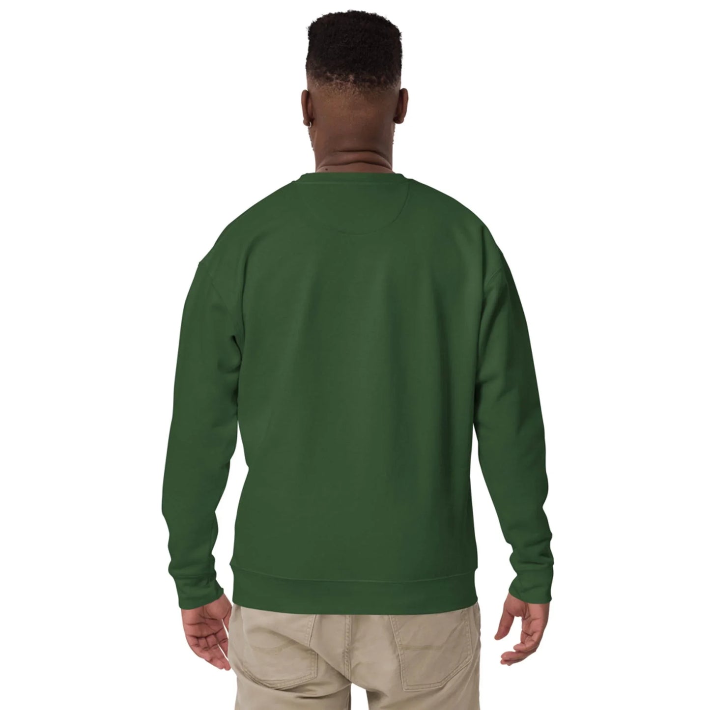 A man stands facing away from the camera, wearing a plain dark green Mambí sweatshirt and light beige pants against a white background.