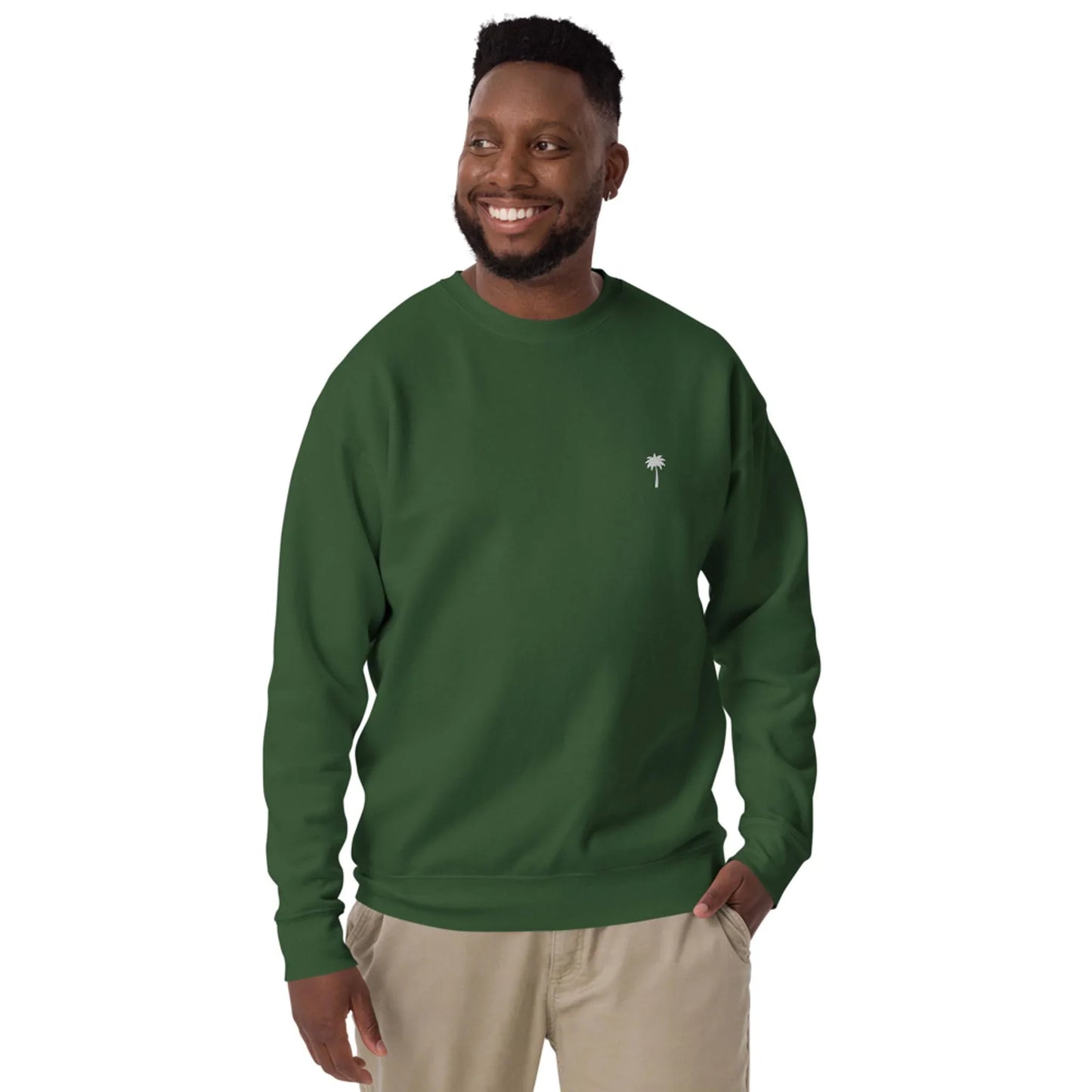 A man smiles while wearing a dark green Mambí sweatshirt with a small white palm tree logo on the left chest, paired with light khaki pants. He stands against a plain white background.