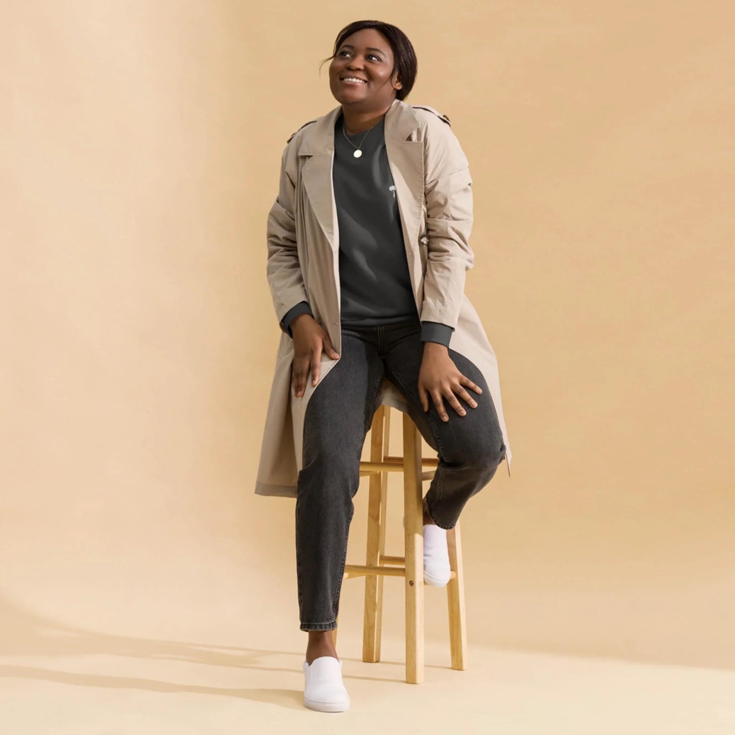 A person wearing a beige Mambí trench coat, dark jeans, and white shoes sits on a wooden stool against a plain beige background, smiling and looking slightly upward.