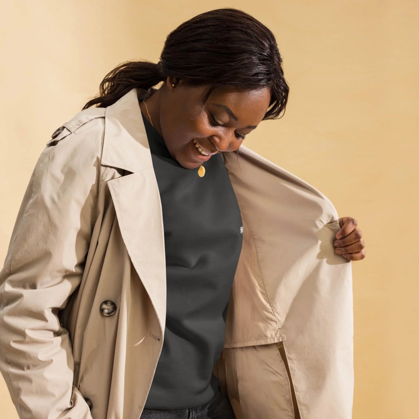 A woman wearing a beige Mambí trench coat and black shirt looks down and smiles, holding open one side of her coat. The background is a solid light beige color.