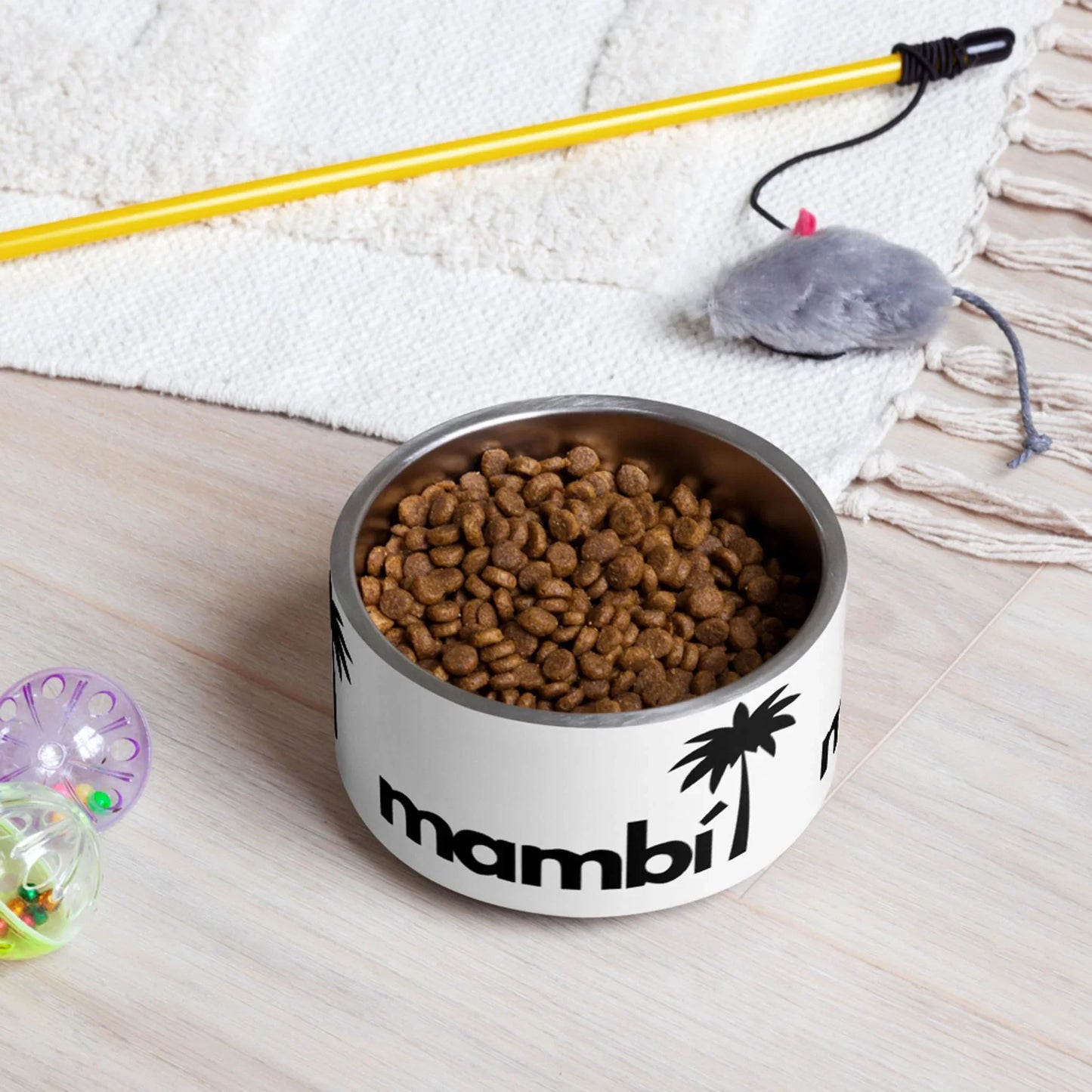 A Printful Simon Stainless Non-Slip Pet Bowl (18oz/32oz), known for being dishwasher-safe, sits on a light wood floor near a cream rug. Cat toys—a plush mouse on a string and a round ball—are nearby.