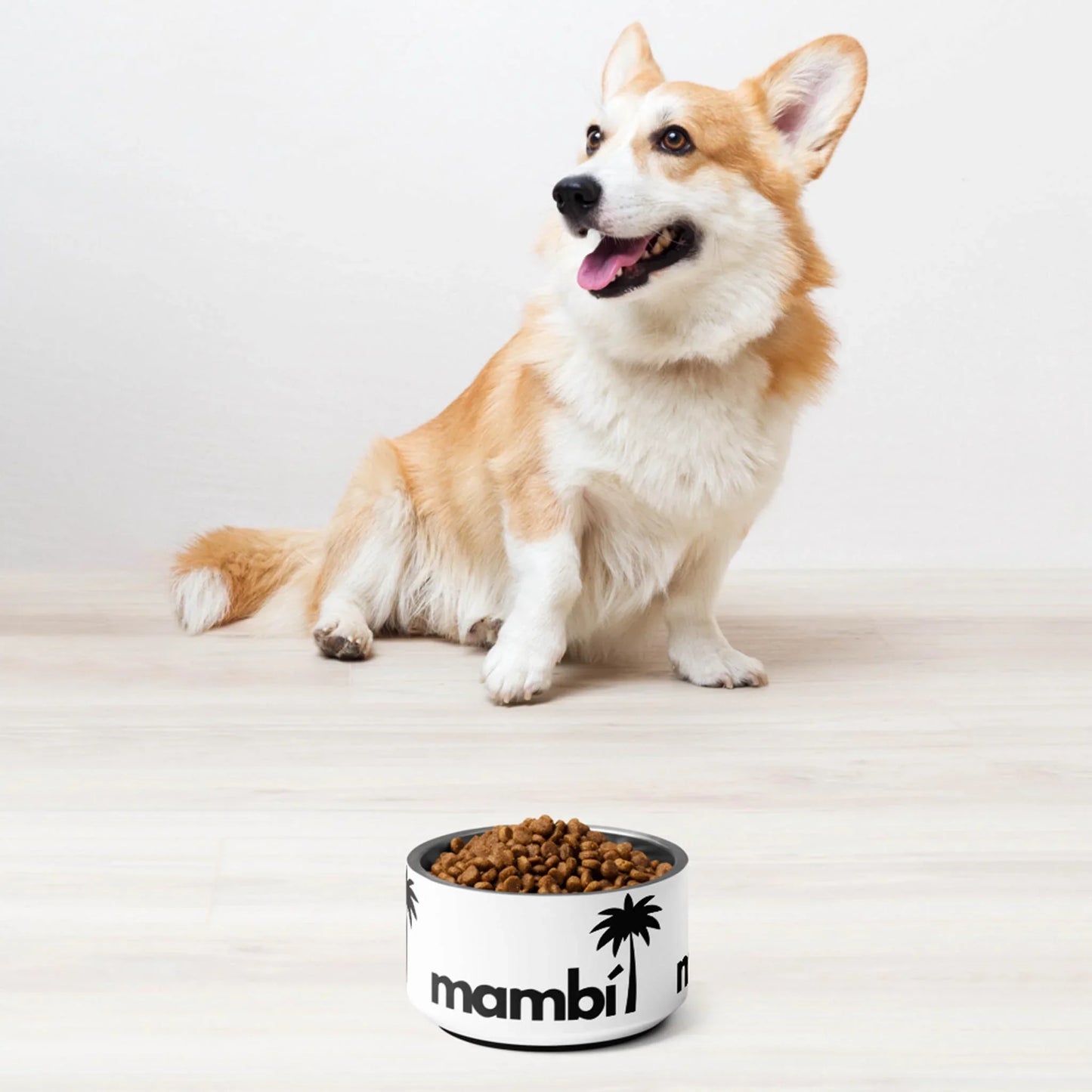 A happy corgi sits on a light wood floor, looking up with its tongue out. In front is Printful’s Simon Stainless Non-Slip Pet Bowl (18oz), dishwasher-safe and filled with kibble, featuring black palm tree graphics and mambi.