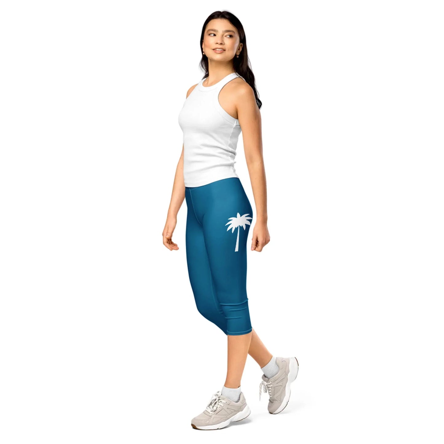 A woman in a white sleeveless top, teal capri leggings with a white palm tree graphic from Mambí, and white sneakers stands and looks to the side, smiling, against a plain white background.
