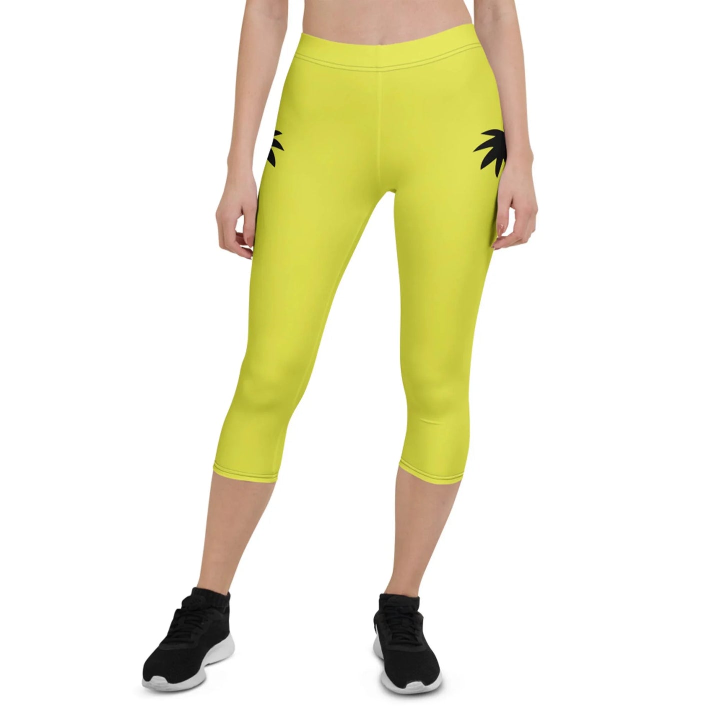 A person wearing bright yellow Mambí capri leggings with black palm leaf prints near the hips, paired with black sneakers. The image shows the lower half of their body against a white background.