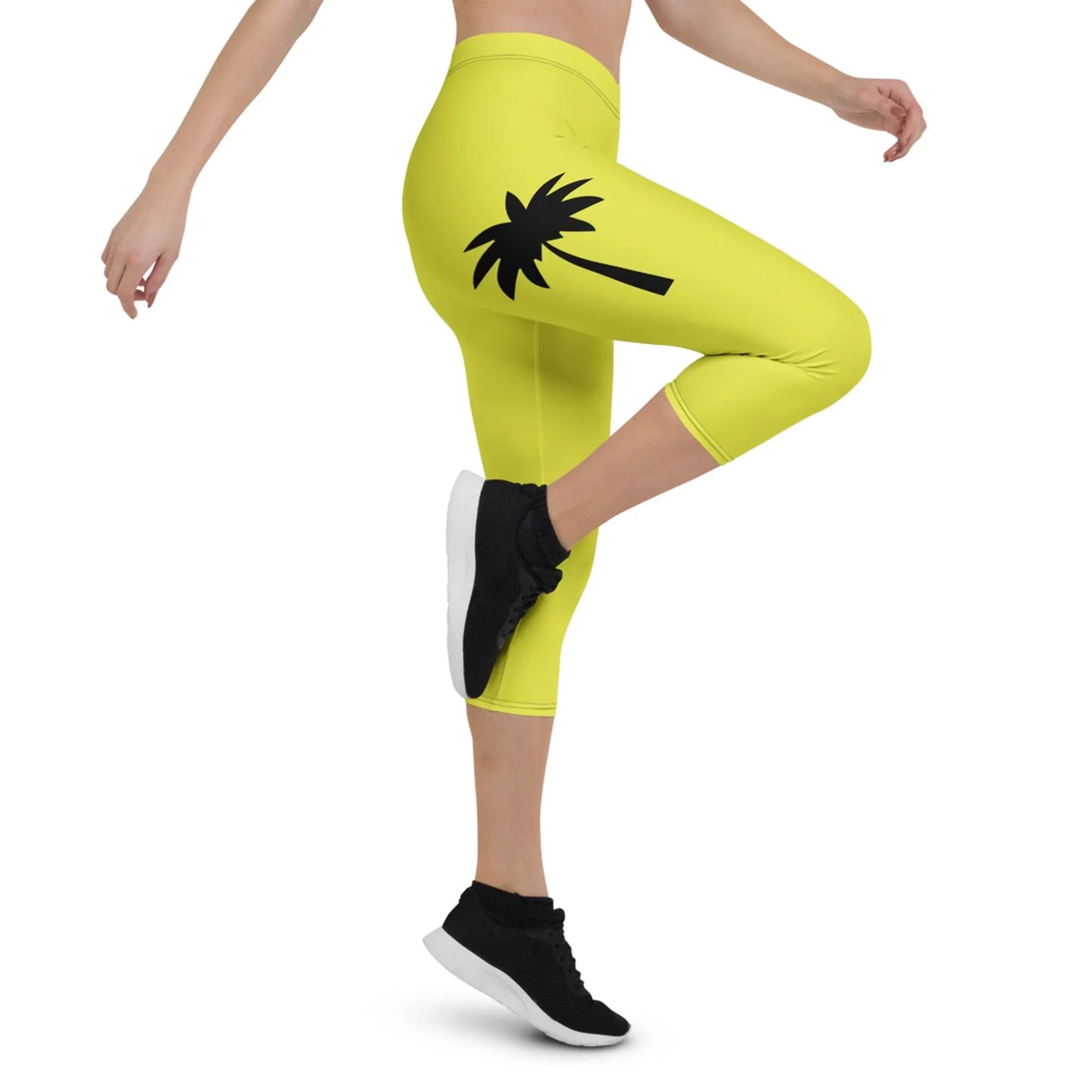 A person wearing bright yellow Mambí capri leggings with a black palm tree graphic on the thigh, black sneakers, and a matching yellow sports top, posing with one leg raised.
