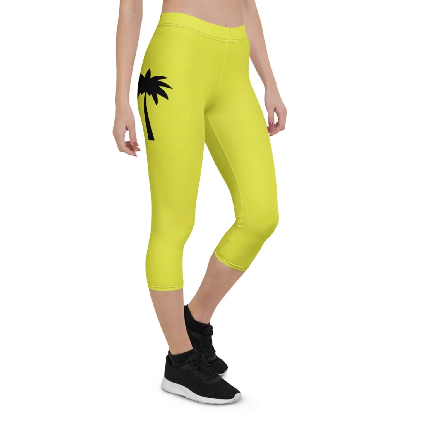 A person wearing bright yellow Mambí capri leggings with a black palm tree graphic on the left hip, black sneakers, and a cropped top stands against a white background.