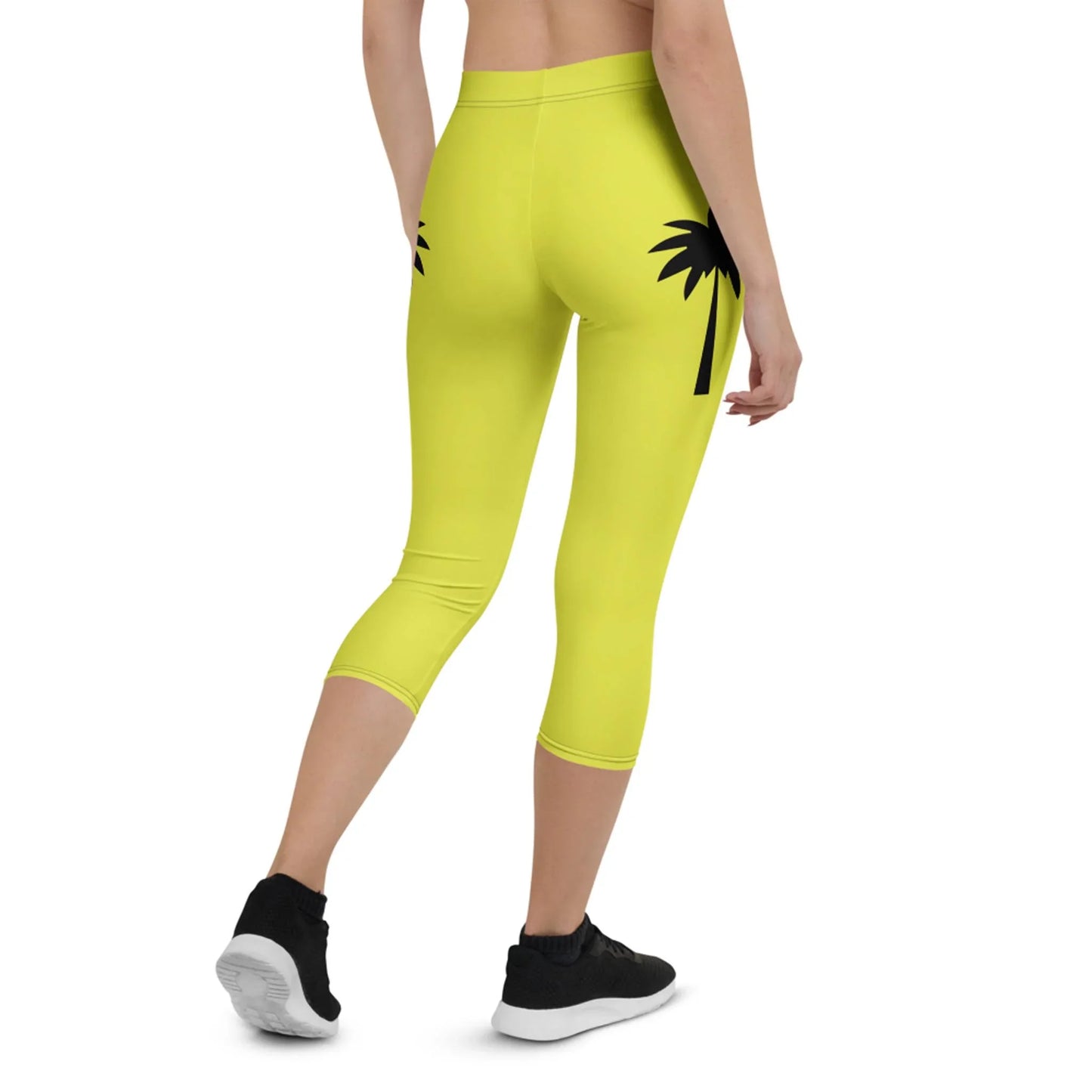 Person wearing bright yellow Mambí capri leggings with a black palm tree graphic on the back, paired with black athletic shoes, shown from behind walking away on a white background.