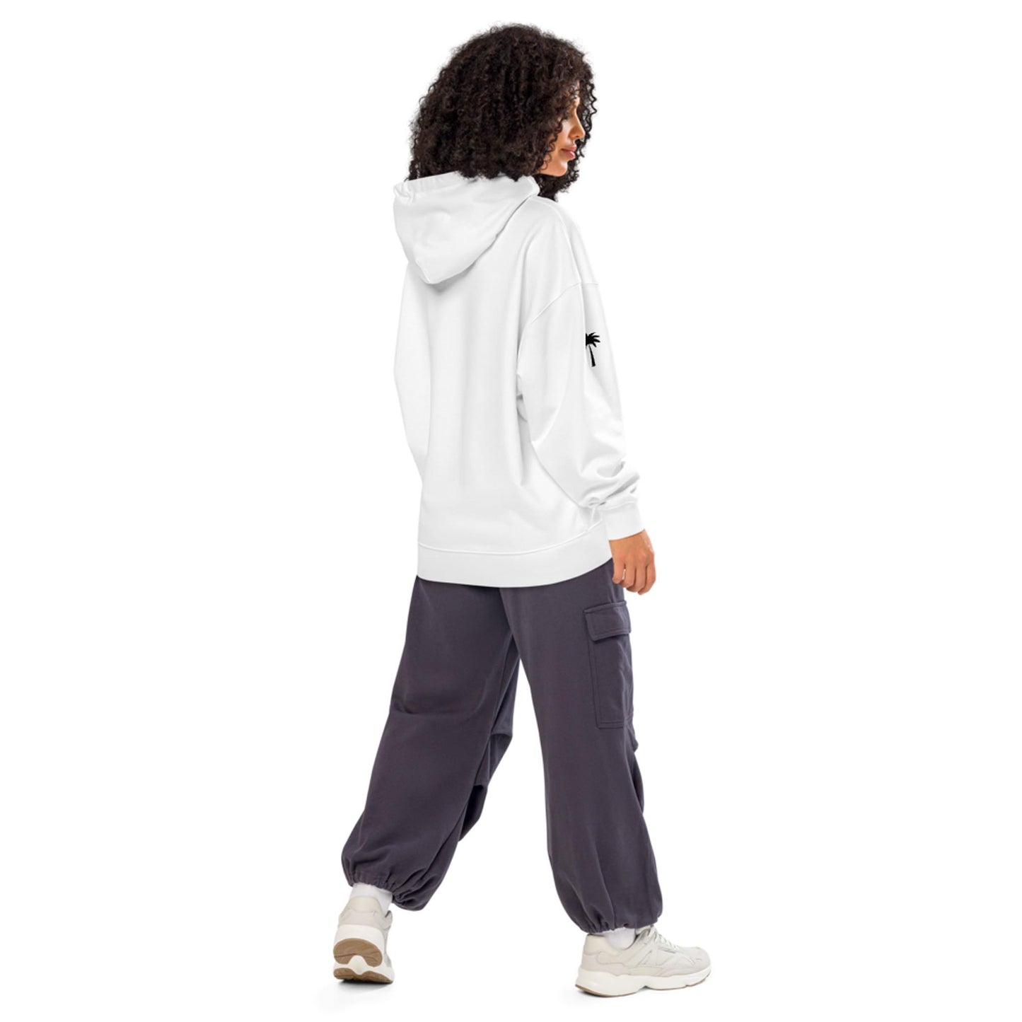 A person with curly hair wears a white Mambí hoodie and dark gray cargo pants with white sneakers, standing and looking to the side against a plain white background.