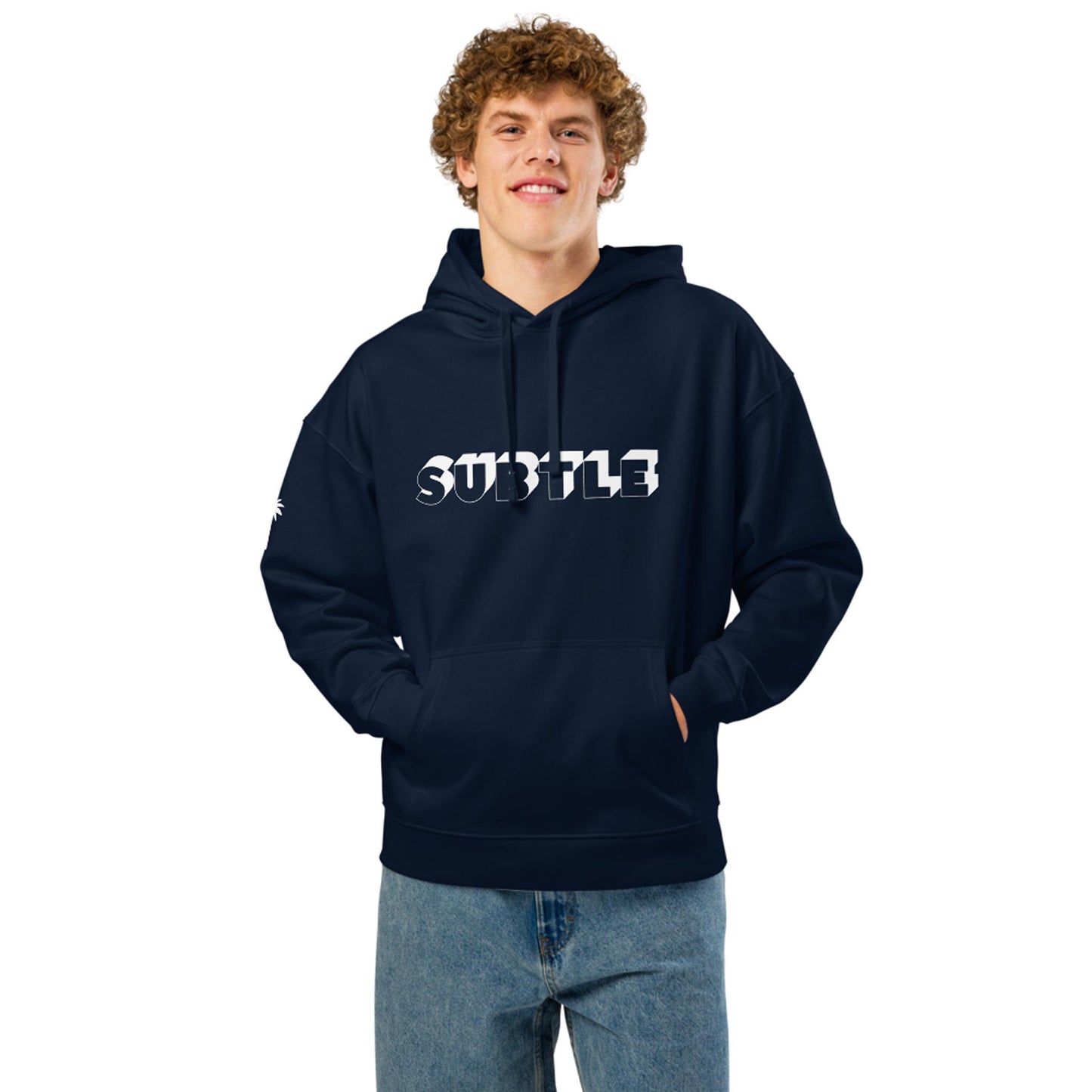A young man with curly hair wears a navy blue Mambí hoodie with the word SUBTLE in bold white letters on the front. He stands with hands in his pocket, smiling at the camera and wearing blue jeans.