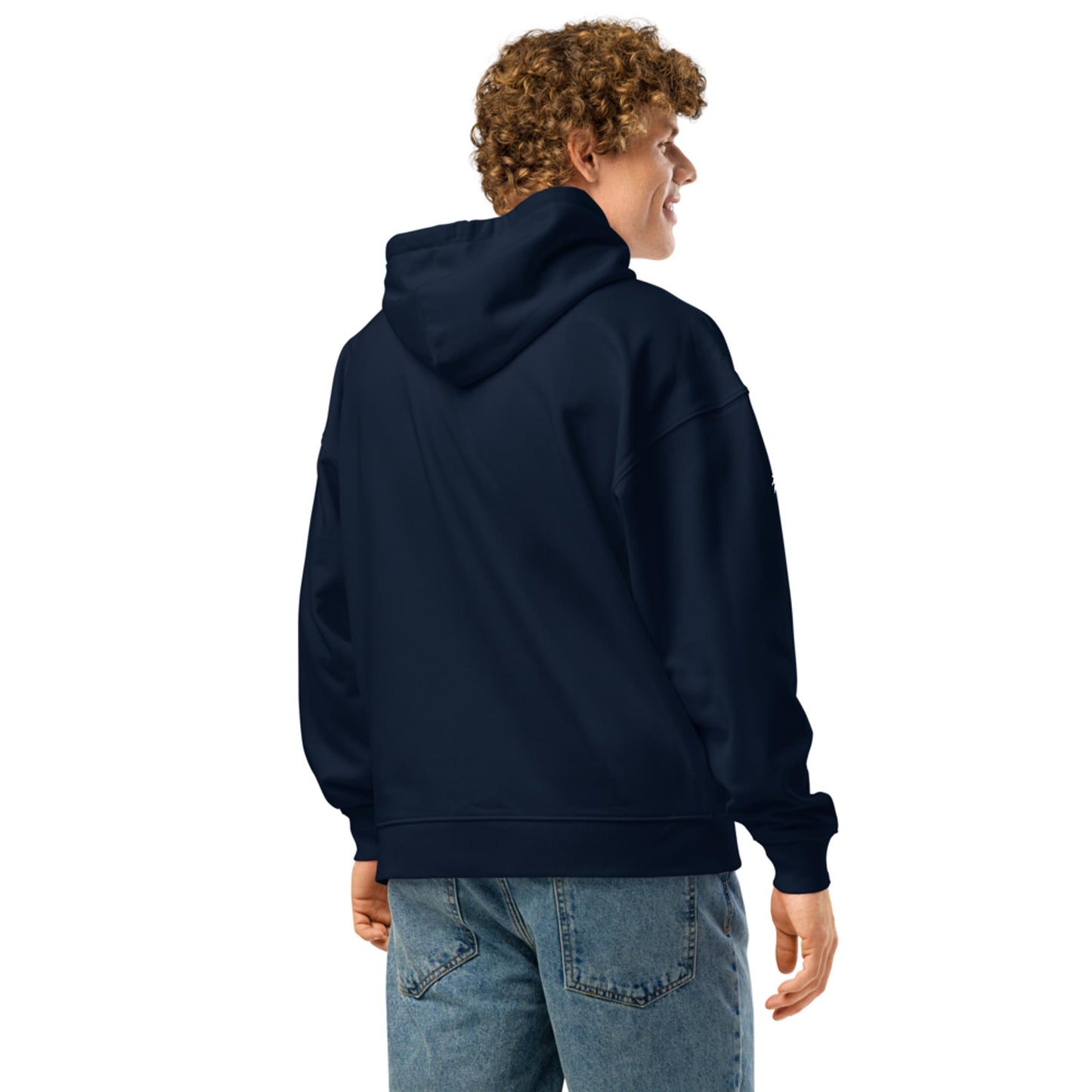 A person with curly hair, wearing a navy blue Mambí hooded sweatshirt and blue jeans, stands facing away and looks to the side against a plain white background.