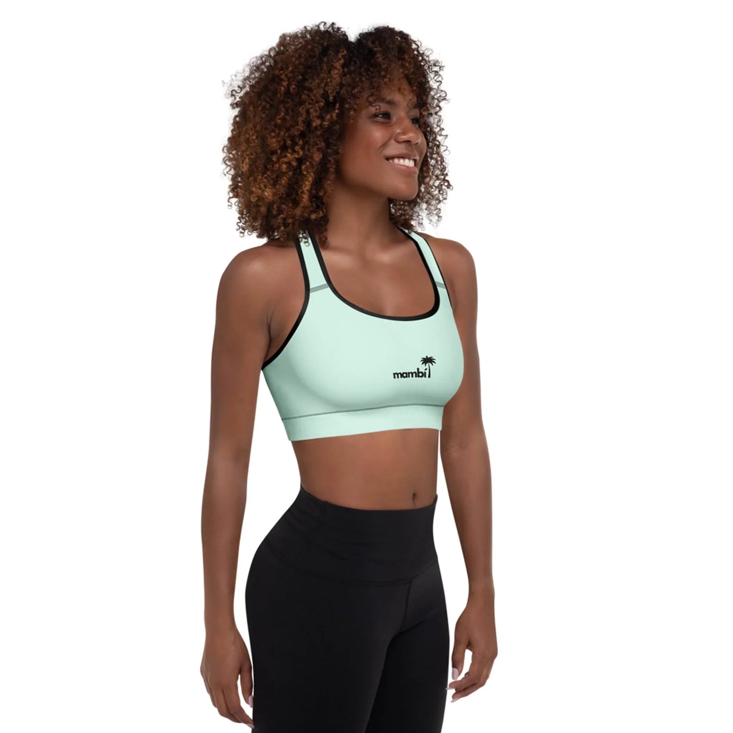 A woman with curly hair smiles, wearing a light blue Mambí sports bra featuring a small palm tree logo and black leggings, standing against a plain white background.