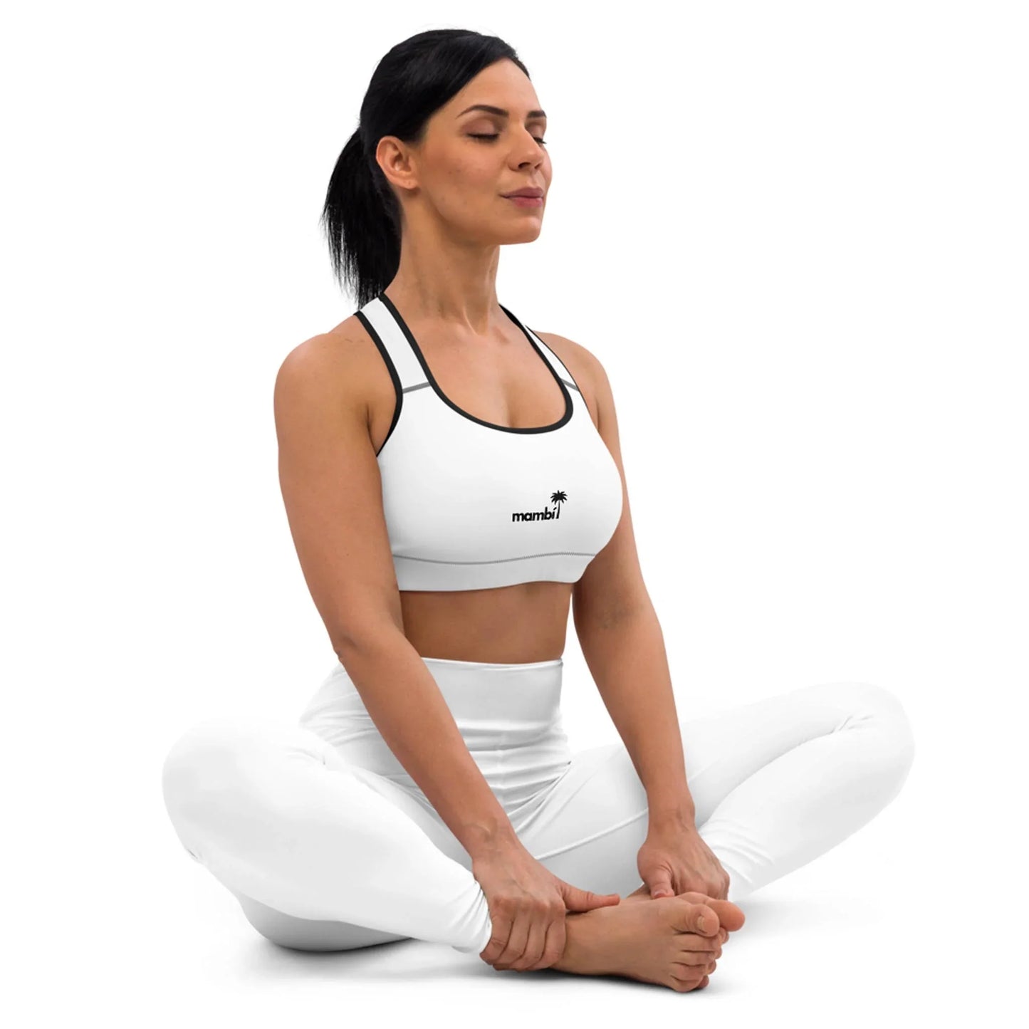 Woman with dark hair in a ponytail sits cross-legged on the floor, wearing a white Mambí sports bra and leggings, eyes closed, appearing calm and meditative against a plain white background.