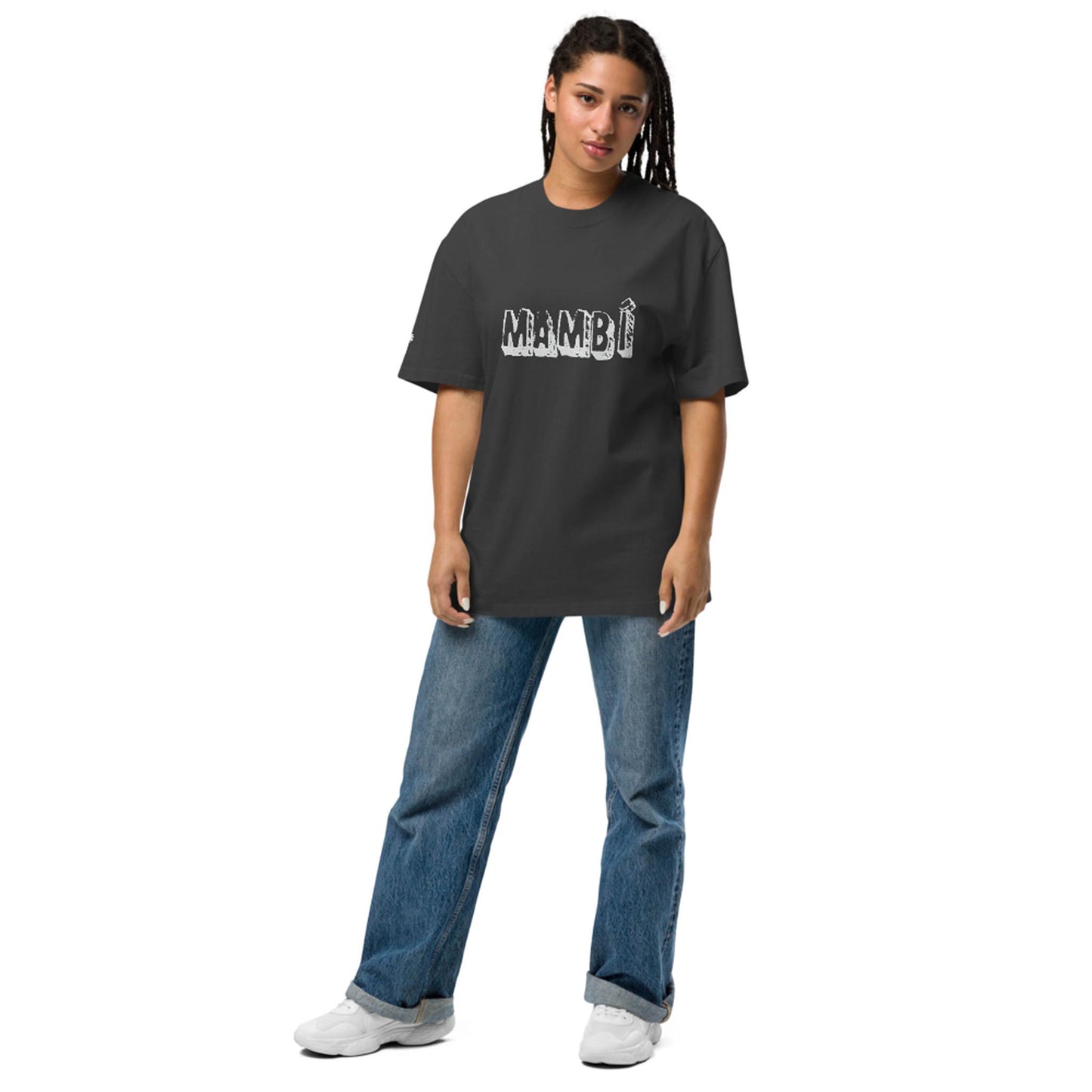 A person with long braided hair wears a loose dark t-shirt featuring the bold, distressed word Mambí, paired with blue jeans and white sneakers, standing against a plain white background.