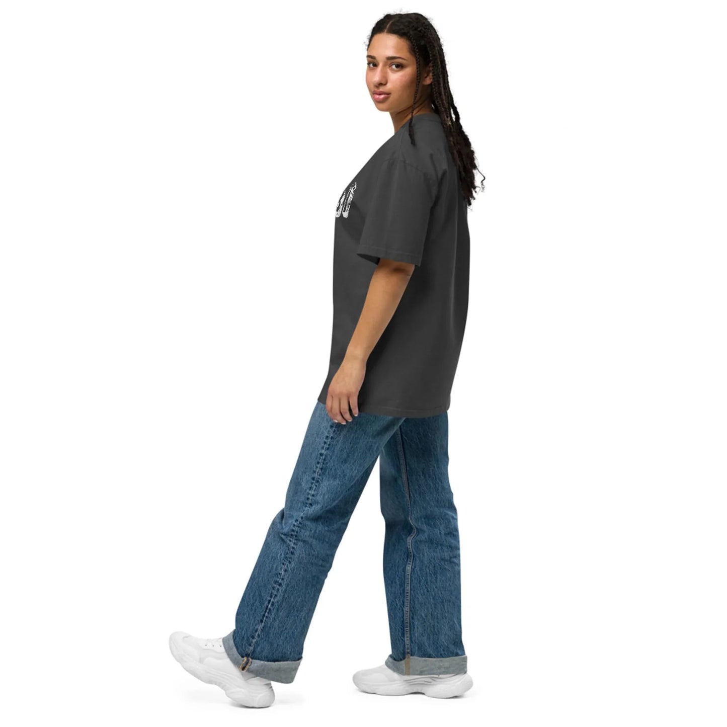 A person with long, dark hair wears a loose dark gray Mambí t-shirt, blue jeans, and white sneakers, standing sideways and looking over their shoulder against a plain white background.