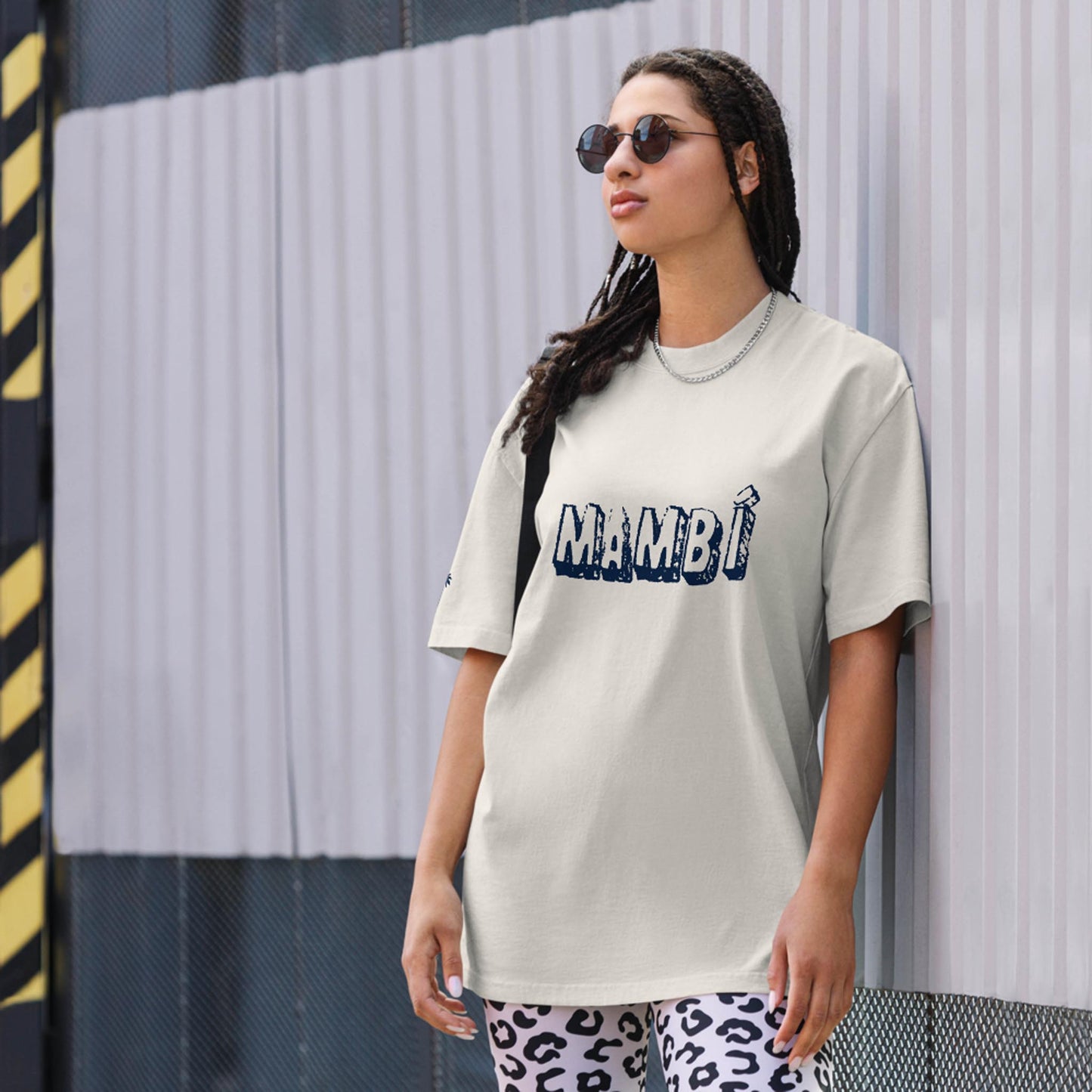 A person with long braided hair and sunglasses leans against a metal wall, wearing a light-colored Mambí t-shirt and animal print leggings.
