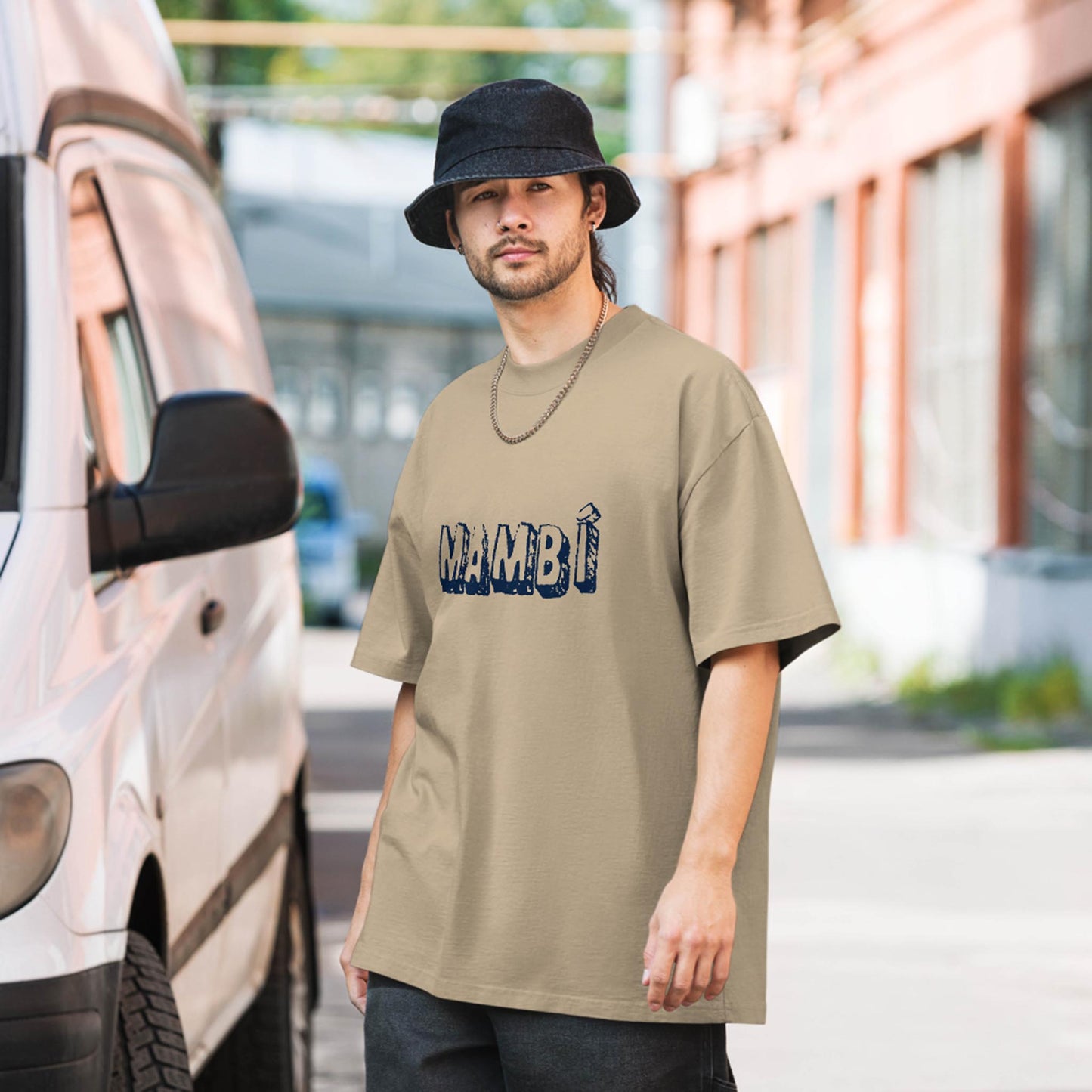 A man wearing a beige Mambí t-shirt, black bucket hat, and dark pants stands next to a white van on a sunlit city street with brick buildings in the background.
