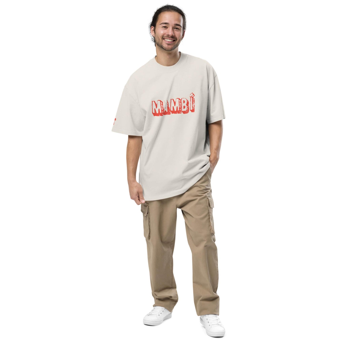A man with long hair in a ponytail smiles, wearing a loose beige Mambí T-shirt with red letters, khaki cargo pants, and white sneakers, standing against a plain white background.