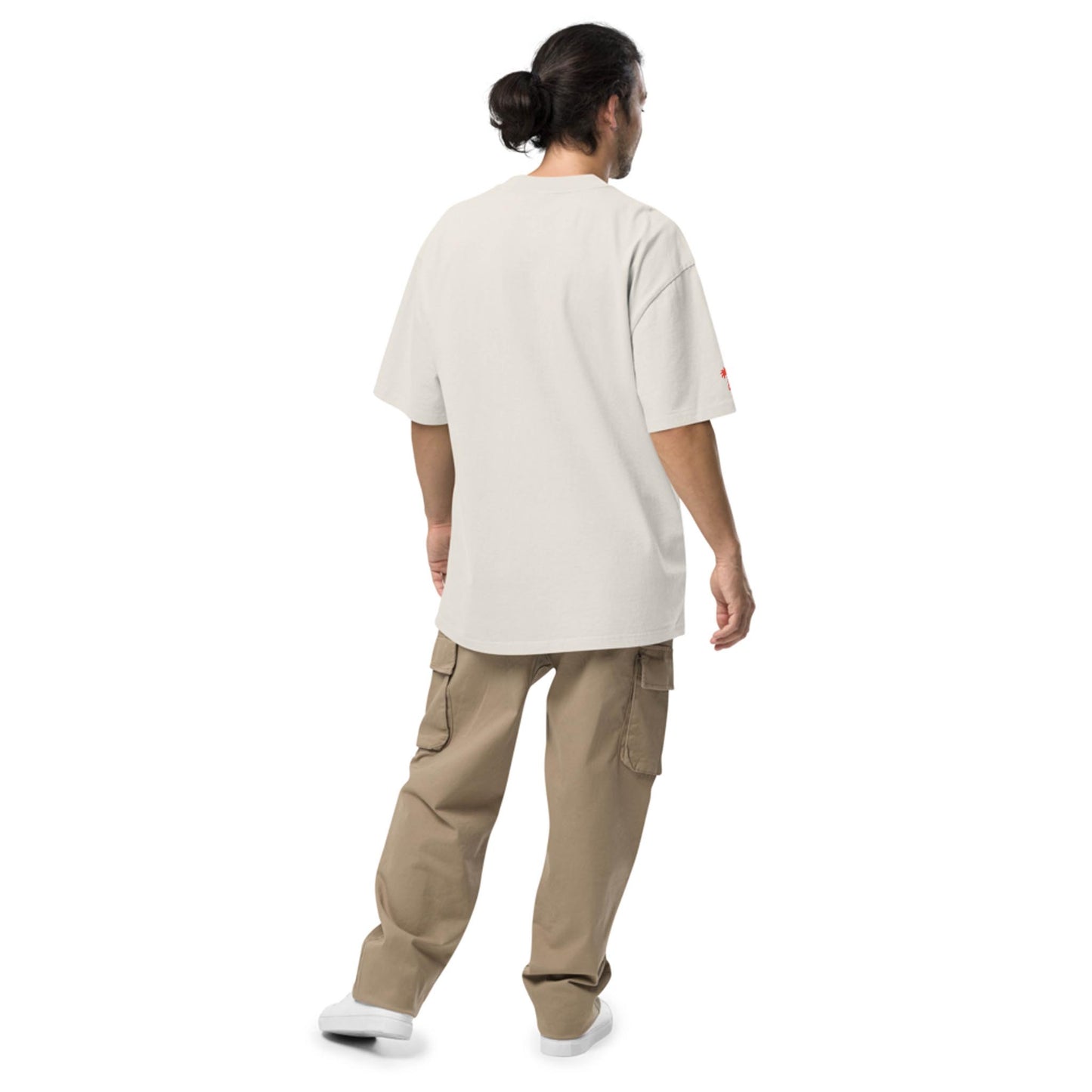 A person with long hair tied in a bun wears a loose white t-shirt, beige cargo pants, and white shoes, standing and facing away from the camera against a plain white background, embodying a relaxed Mambí-inspired style.
