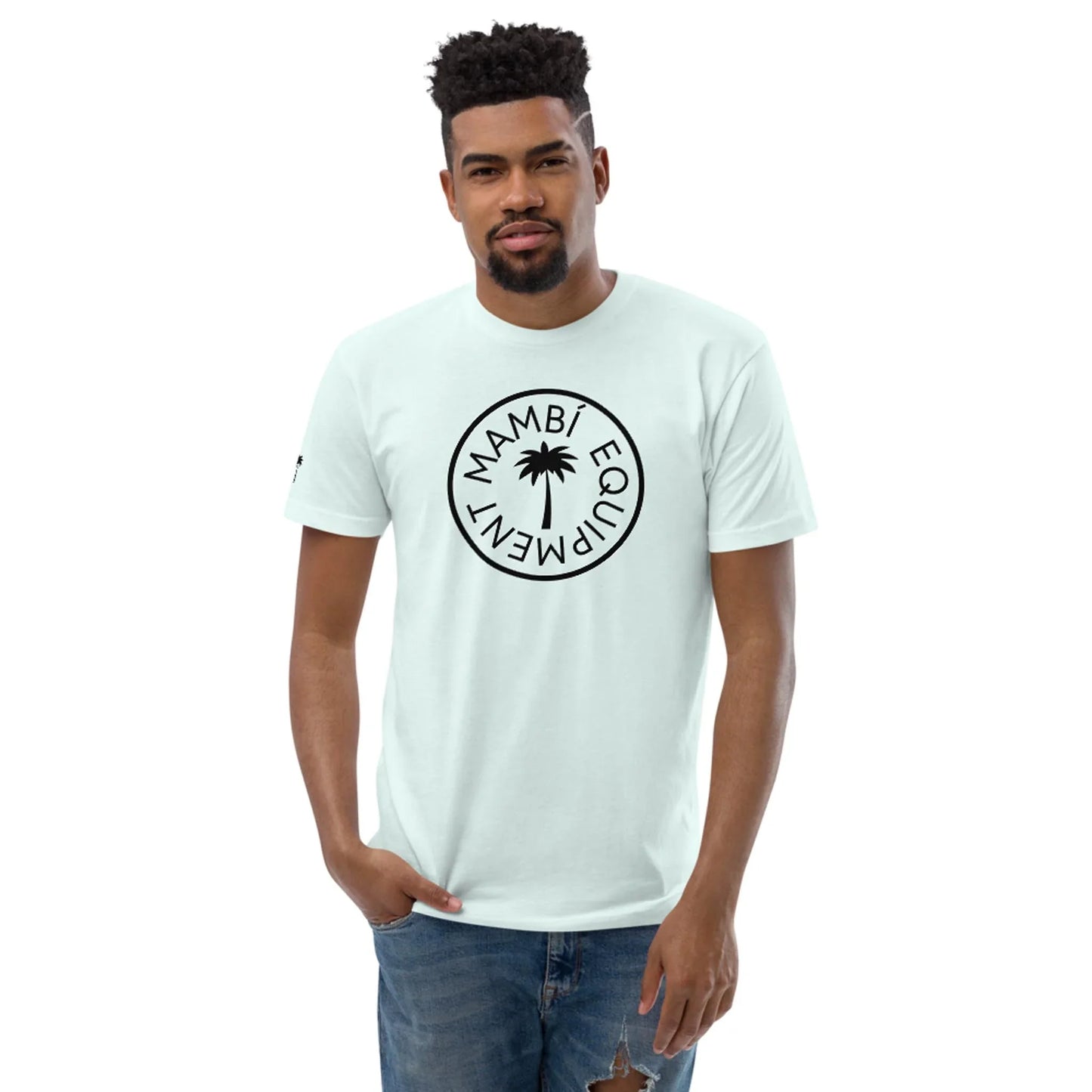 A man with short curly hair and a beard wears a light blue Mambí T-shirt featuring a circular logo and palm tree design. He stands with one hand in his pocket, dressed in distressed blue jeans.