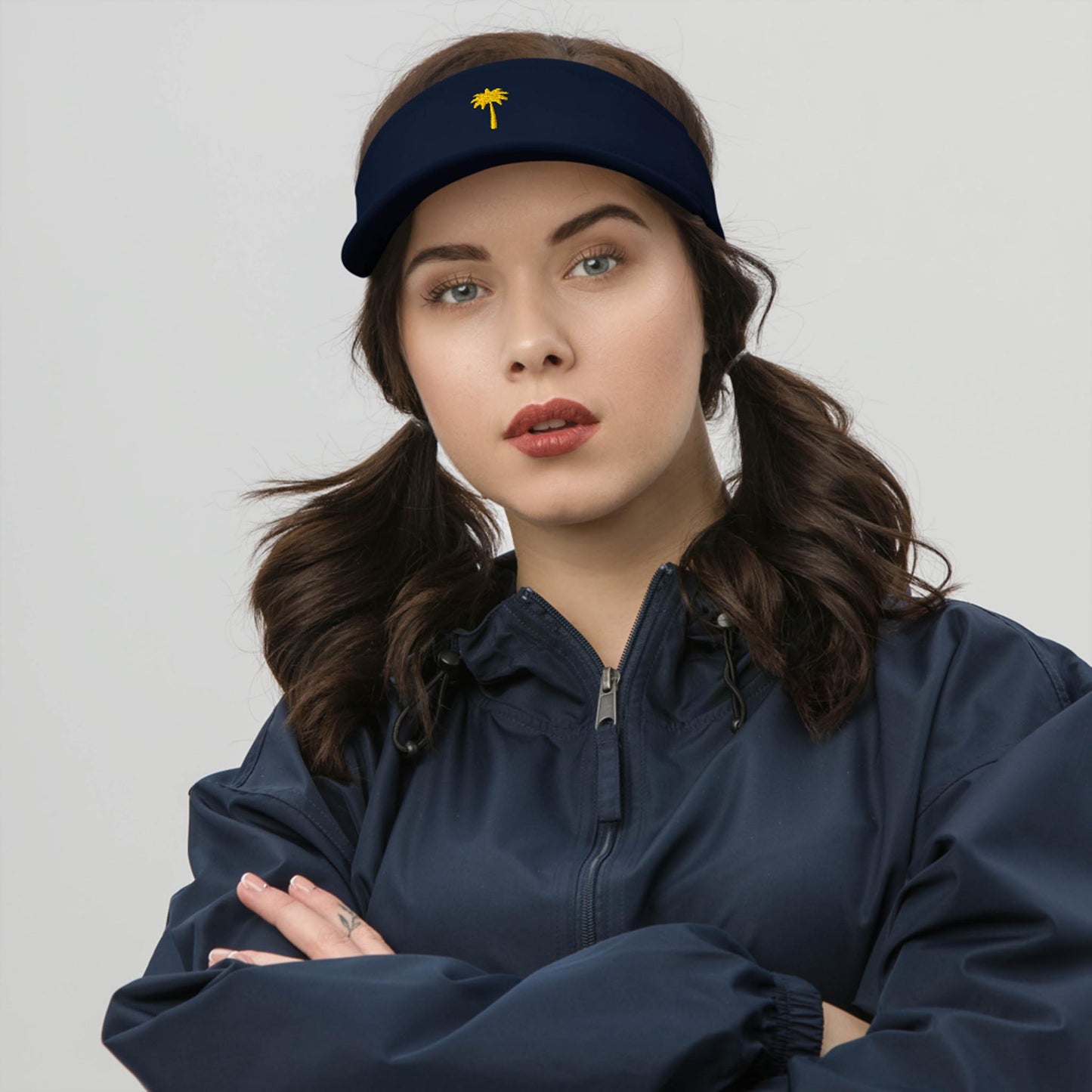 A woman with dark brown hair in pigtails wears a navy visor featuring a small yellow palm tree emblem and a matching Mambí navy jacket, standing with folded arms against a plain light background.