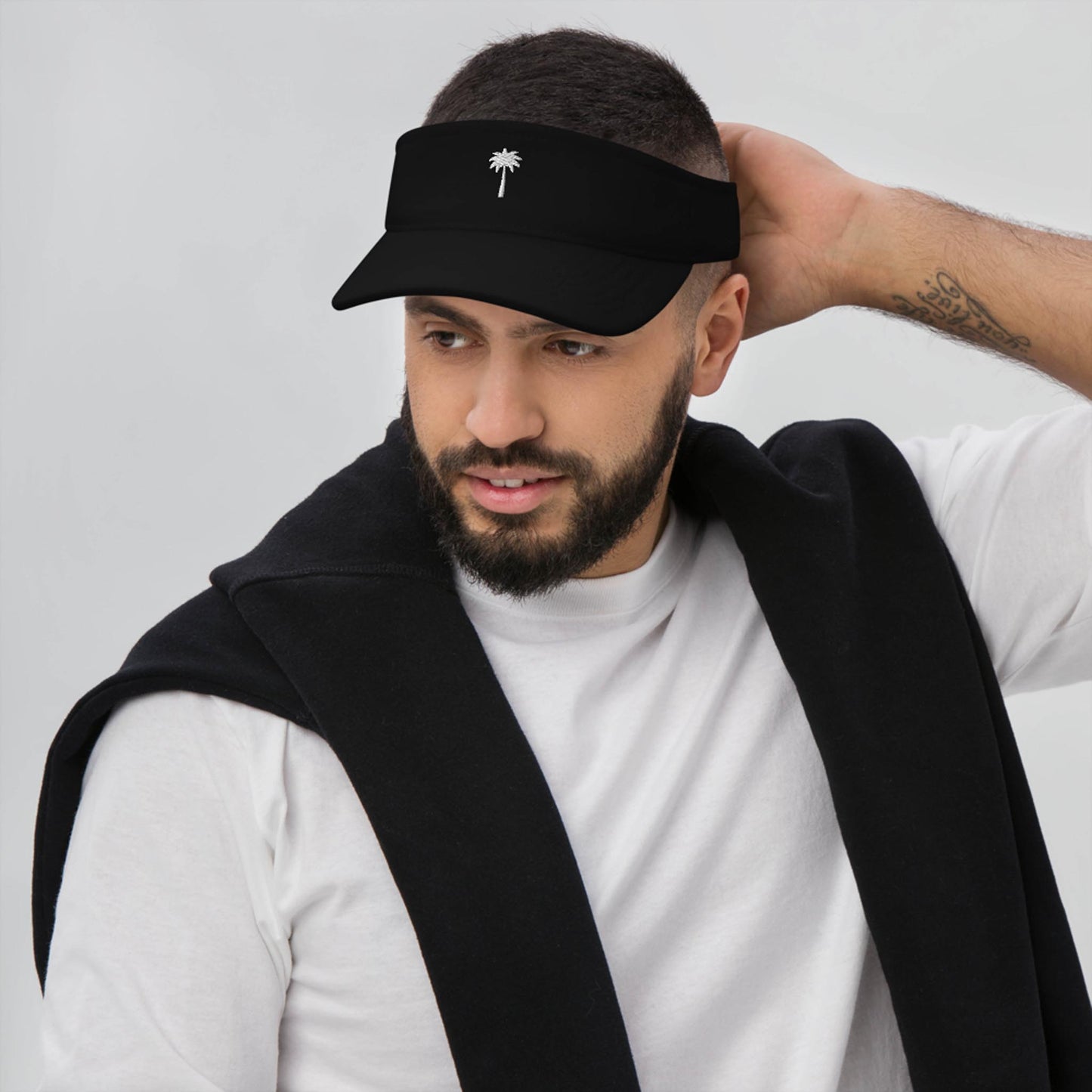 A man with a short beard and mustache wears a black Mambí visor with a white palm tree logo, a white shirt, and a black sweater draped over his shoulders, looking to the side and touching the back of his head.
