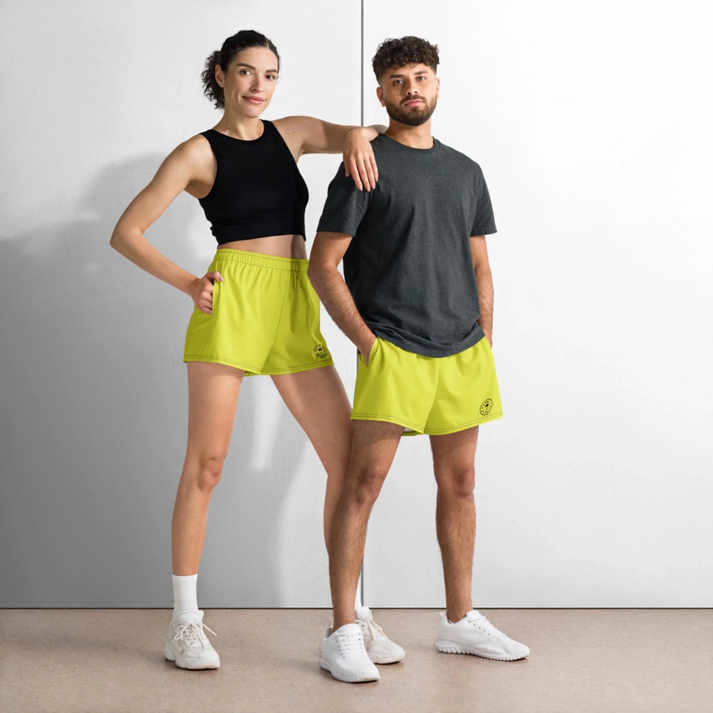 Two people stand confidently side by side in neon yellow Mambí athletic shorts and white sneakers. The woman wears a black sleeveless crop top; the man sports a dark gray t-shirt. They pose against a minimalist white wall.