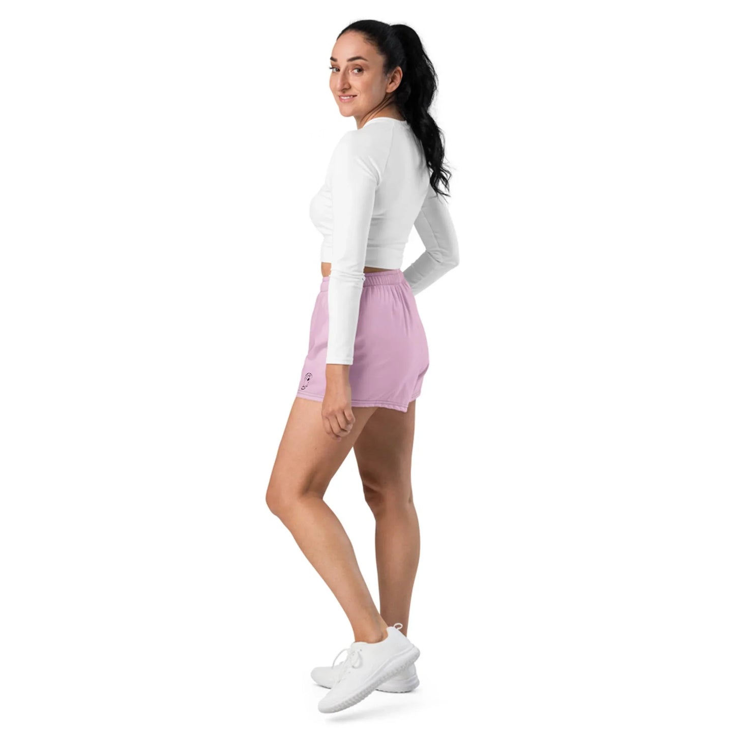 A woman with long dark hair in a ponytail wears a white long-sleeve top, light pink shorts, and white sneakers. She stands sideways, looking back and smiling against a white background, exuding effortless Mambí style.